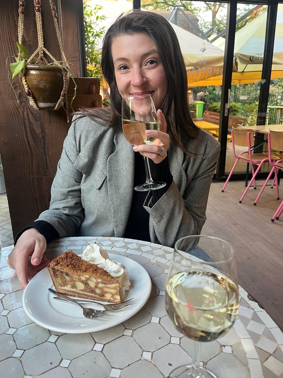 A woman with dark hair smiling and holding a glass of white wine, sitting at a table with a slice of apple pie topped with whipped cream and a glass of white wine, in a cafe with outdoor seating visible through the window.