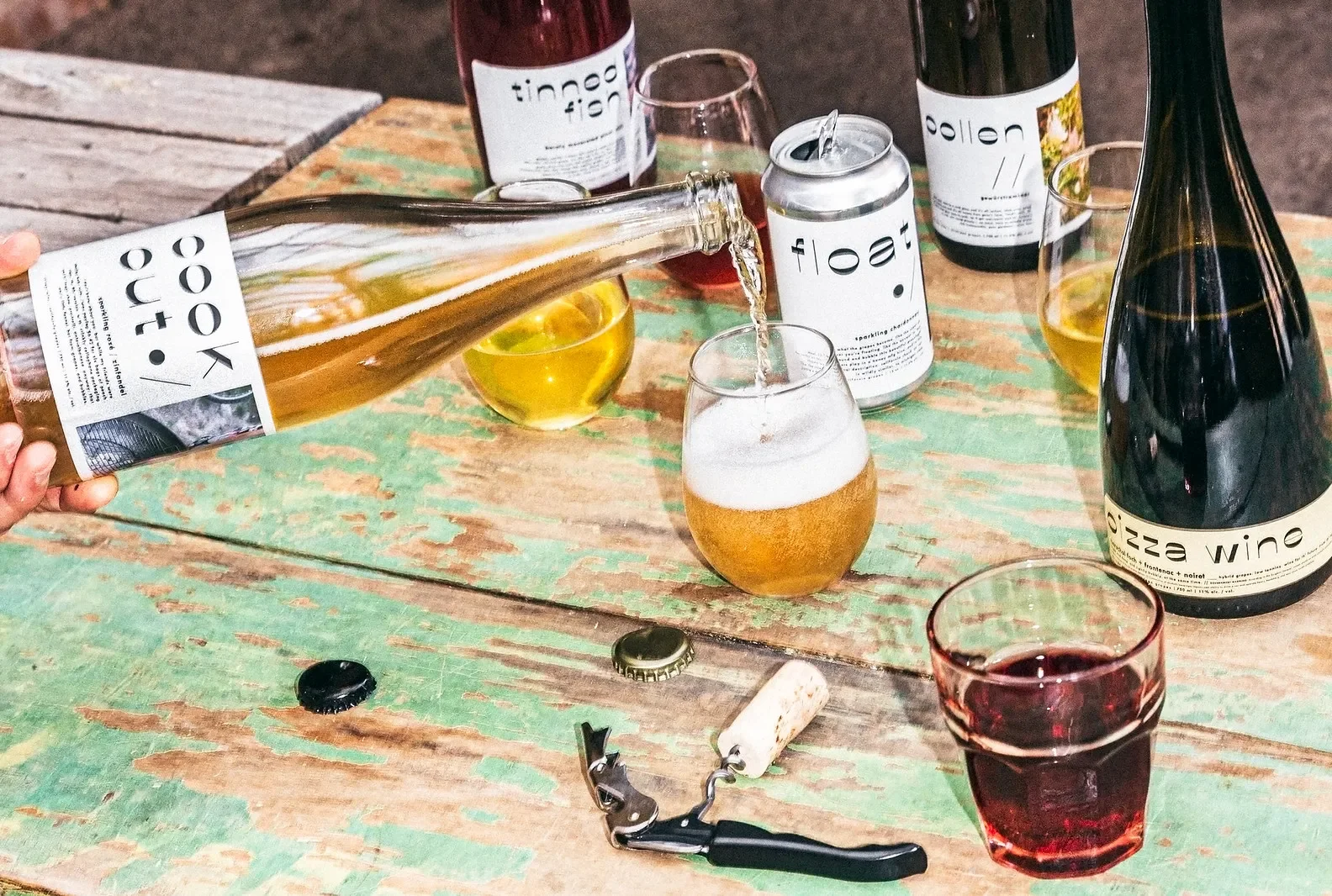 A wooden table with various wine bottles, glasses filled with wine, a beer bottle, and a glass of beer. Someone is pouring beer into a glass from a bottle labeled 'look out!'. There are some corks, a bottle opener, and a crown cap on the table.