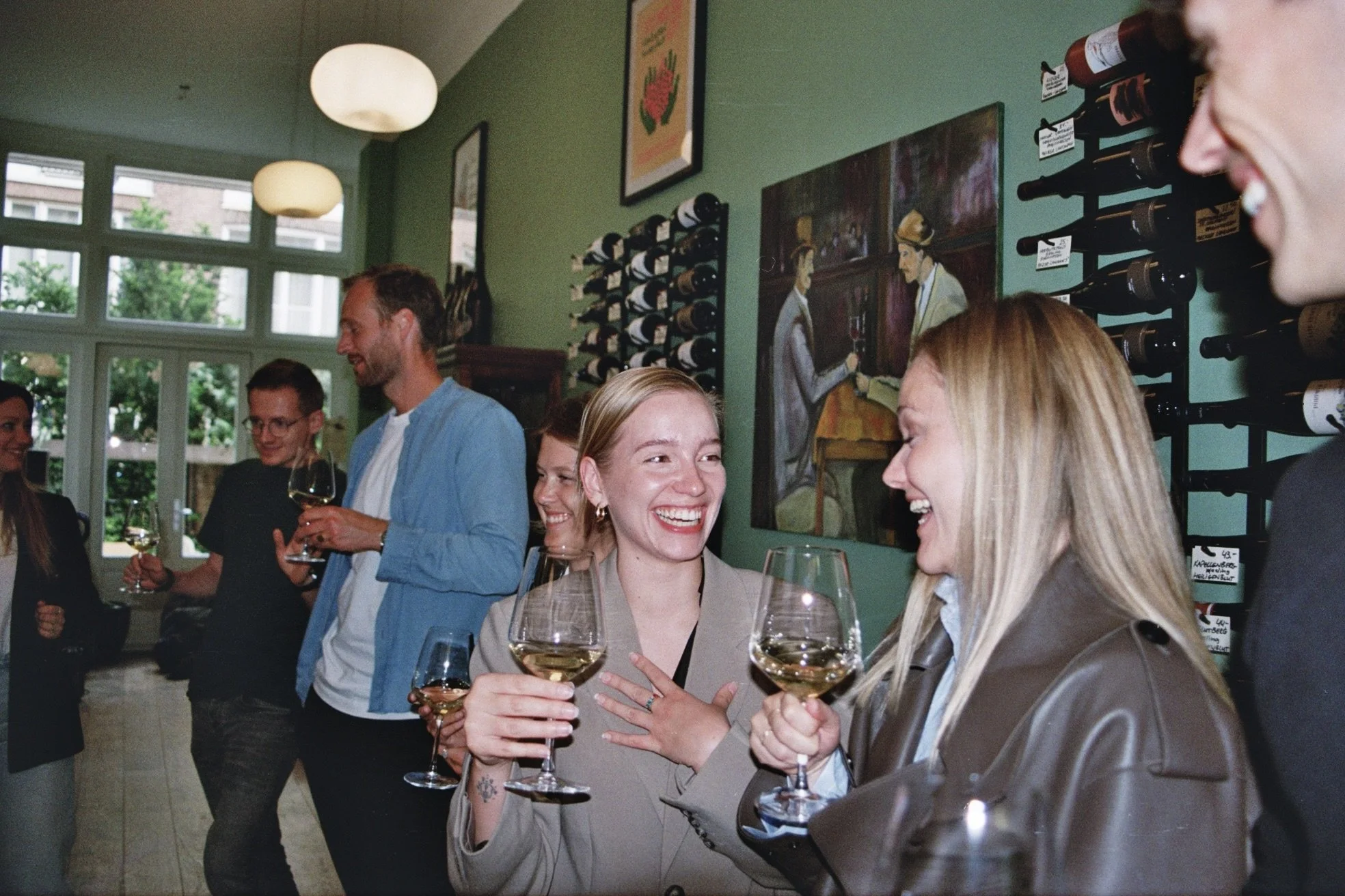People smiling and talking while holding glasses of white wine at a social gathering in a cafe or wine bar.