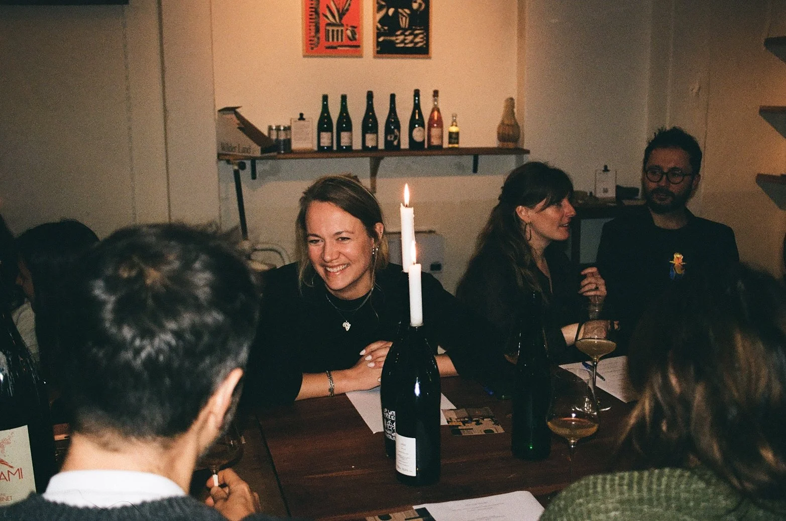 People gathered around a table at a celebration; one woman in the center is smiling and wearing a black outfit with a necklace, surrounded by others holding drinks, with a candle on a wine bottle on the table and bottles of wine and artwork on the wall in the background.