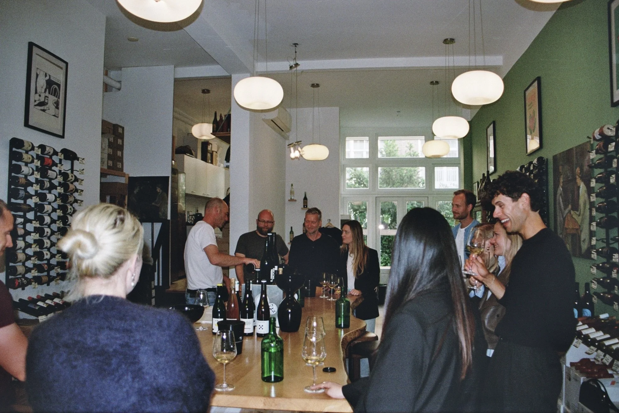 Group of people gathered in a wine tasting event inside a modern, brightly lit room with wine bottles on display and a large window in the background.