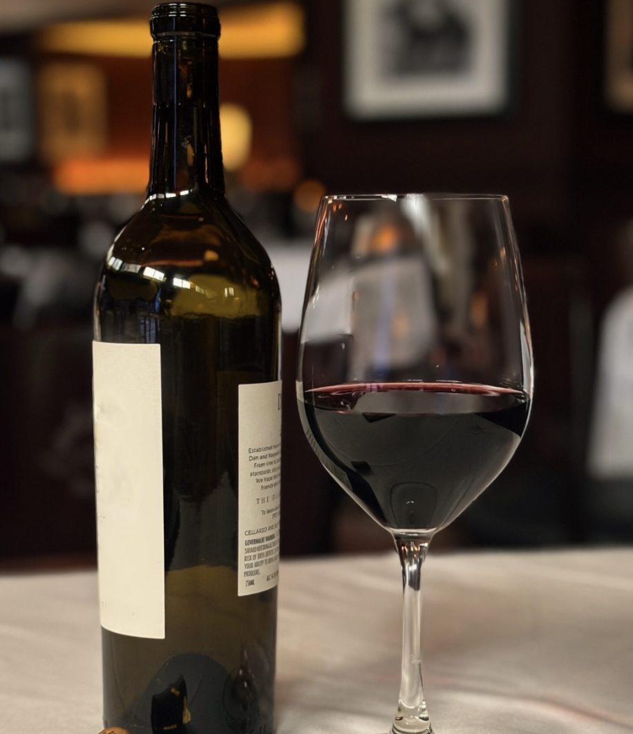 A bottle of red wine and a wine glass filled with red wine on a table in a dimly lit setting.