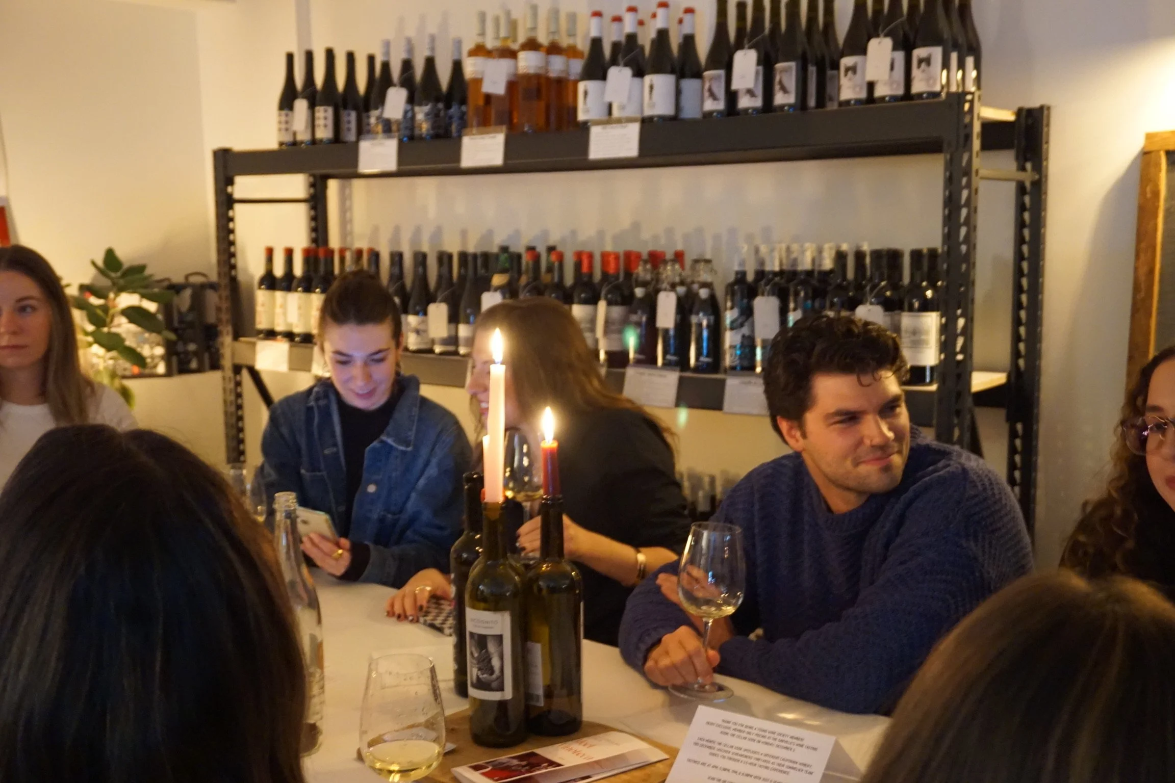 People enjoying a dinner party in a cozy restaurant, with wine bottles on shelves in the background and candles on the table.