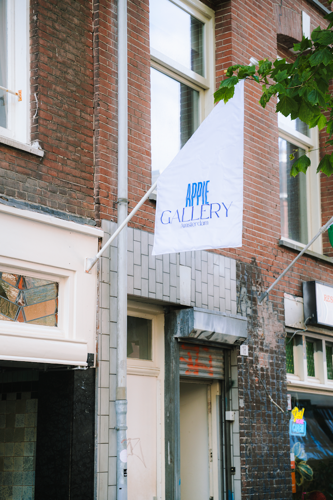 The image shows the exterior of a building with a white flag that reads "APPLE GALLERY Amsterdam" hanging from a pole. The building has red brick walls, white window frames, and a closed metal shutter with graffiti. There are some trees with green leaves partially visible.