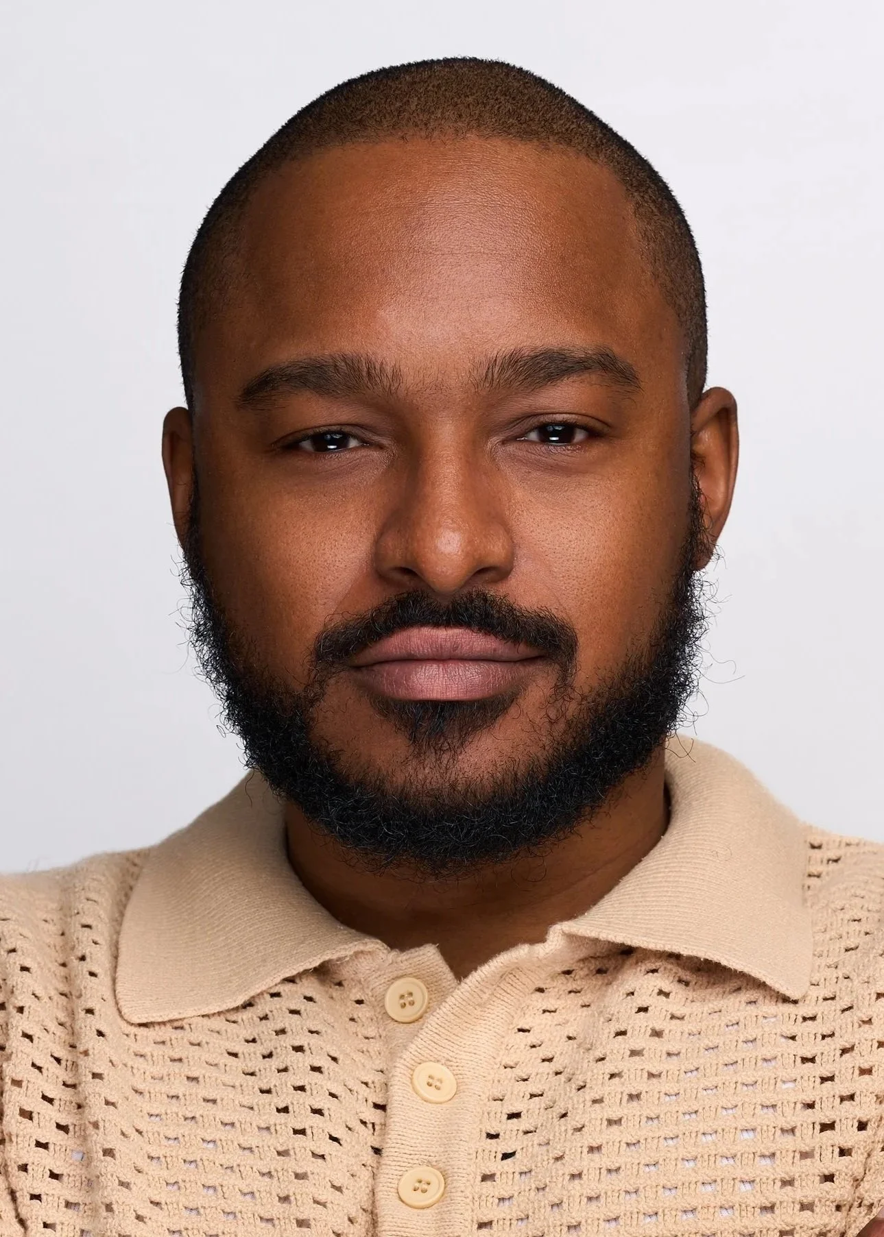 A smiling man with short black hair, a beard, wearing a black polo shirt, and a gold chain, standing against a plain light gray background.