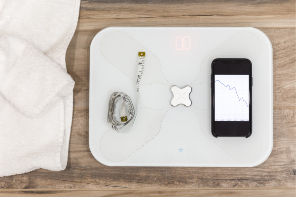 Weegschaal met meetlint en een smartphone met een grafiek, naast een wit badhanddoek op een houten vloer.