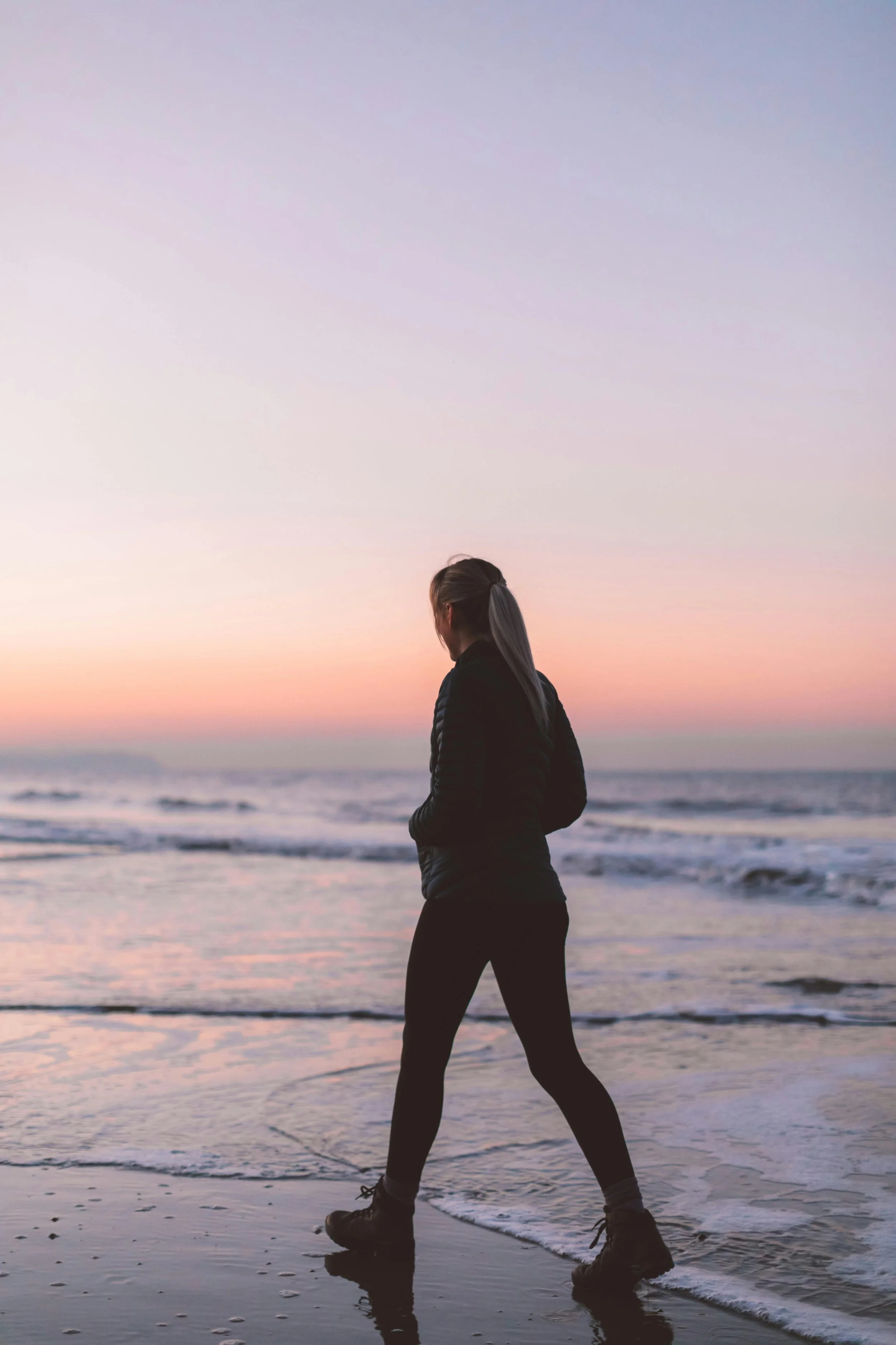 Vrouw in zwart jack en legging wandelt op het strand bij sunset, zicht vanaf achterkant.