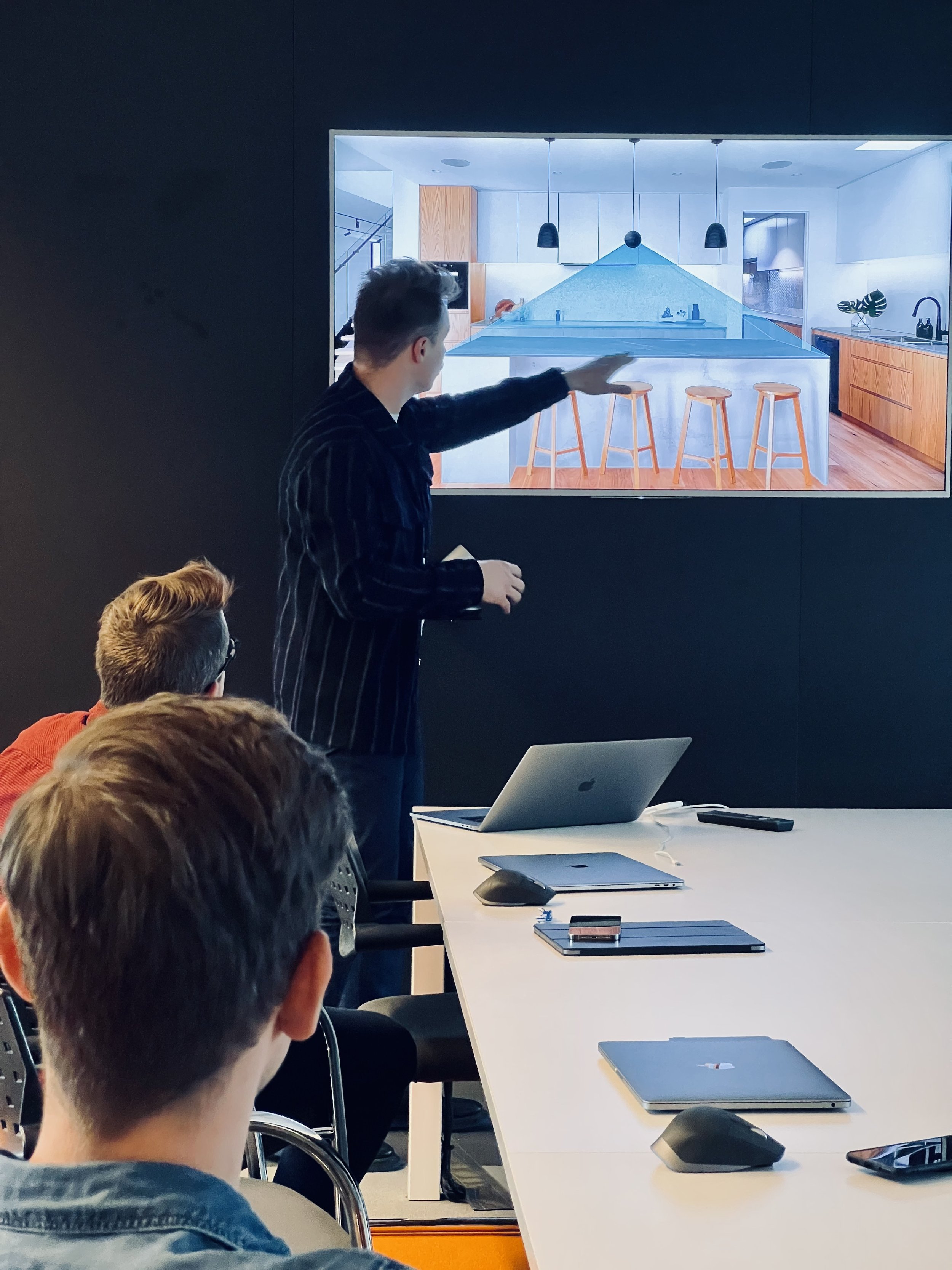 A person giving a presentation pointing at a screen showing a modern kitchen design with bar stools. Several attendees are seated at a conference table with laptops and notepads.
