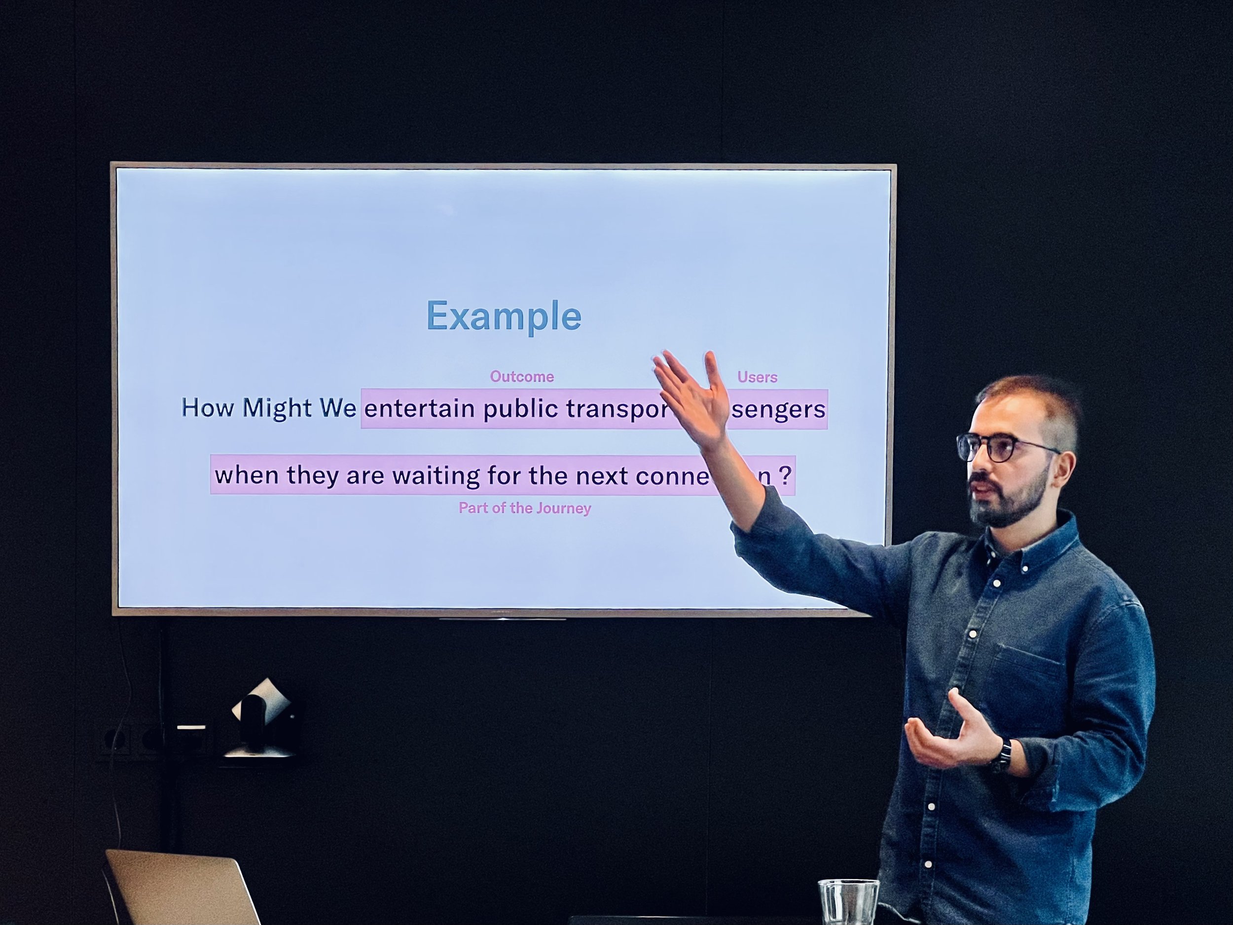 Man giving a presentation with a screen displaying a design thinking question about public transportation. He gestures toward the screen which shows text: "Example: How Might We entertain public transport passengers when they are waiting for the next