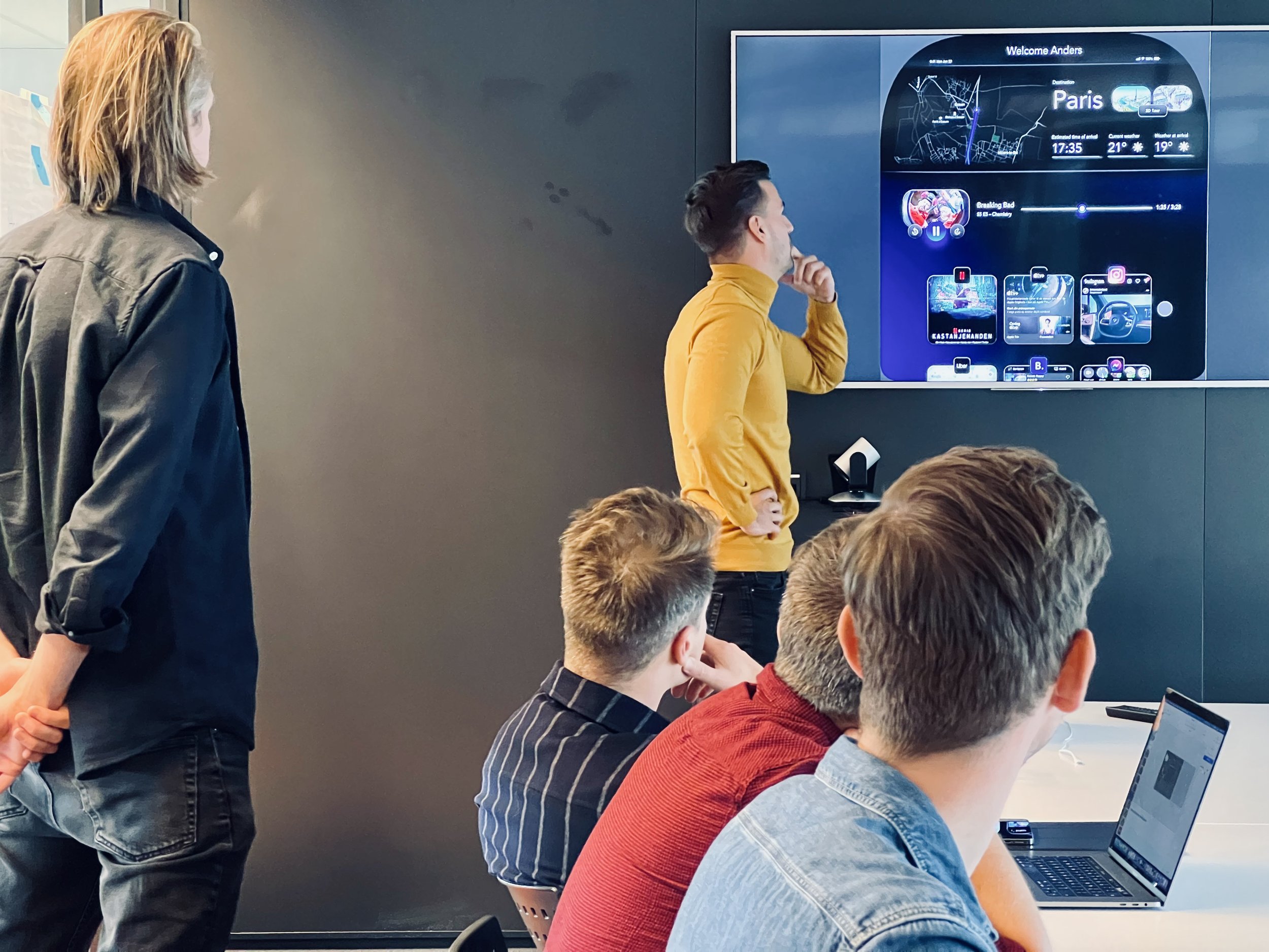 Group of people in a meeting room looking at a large screen displaying a user interface with travel information and media options.