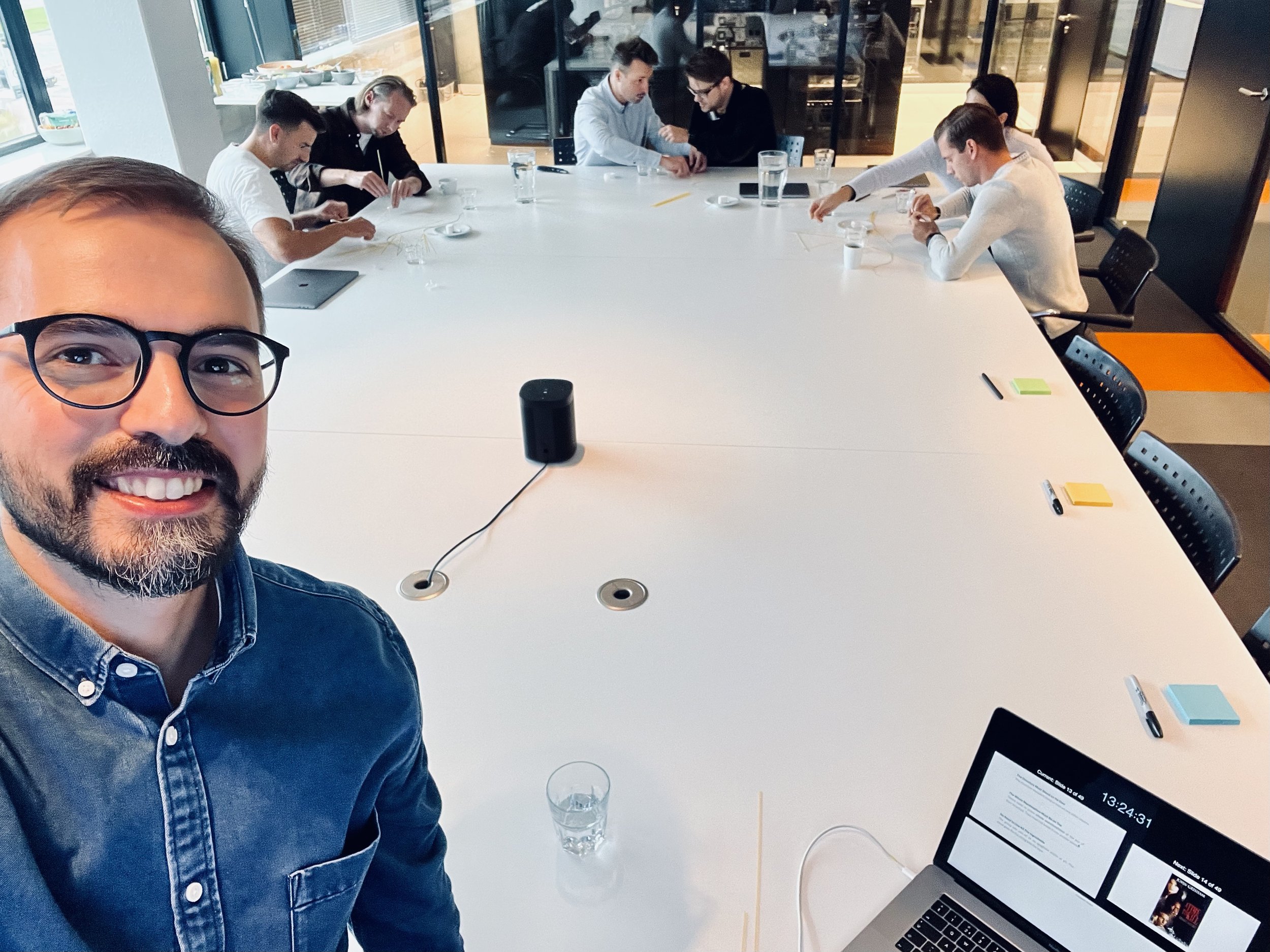 Group meeting in an office with a person taking a selfie and others working at a conference table.
