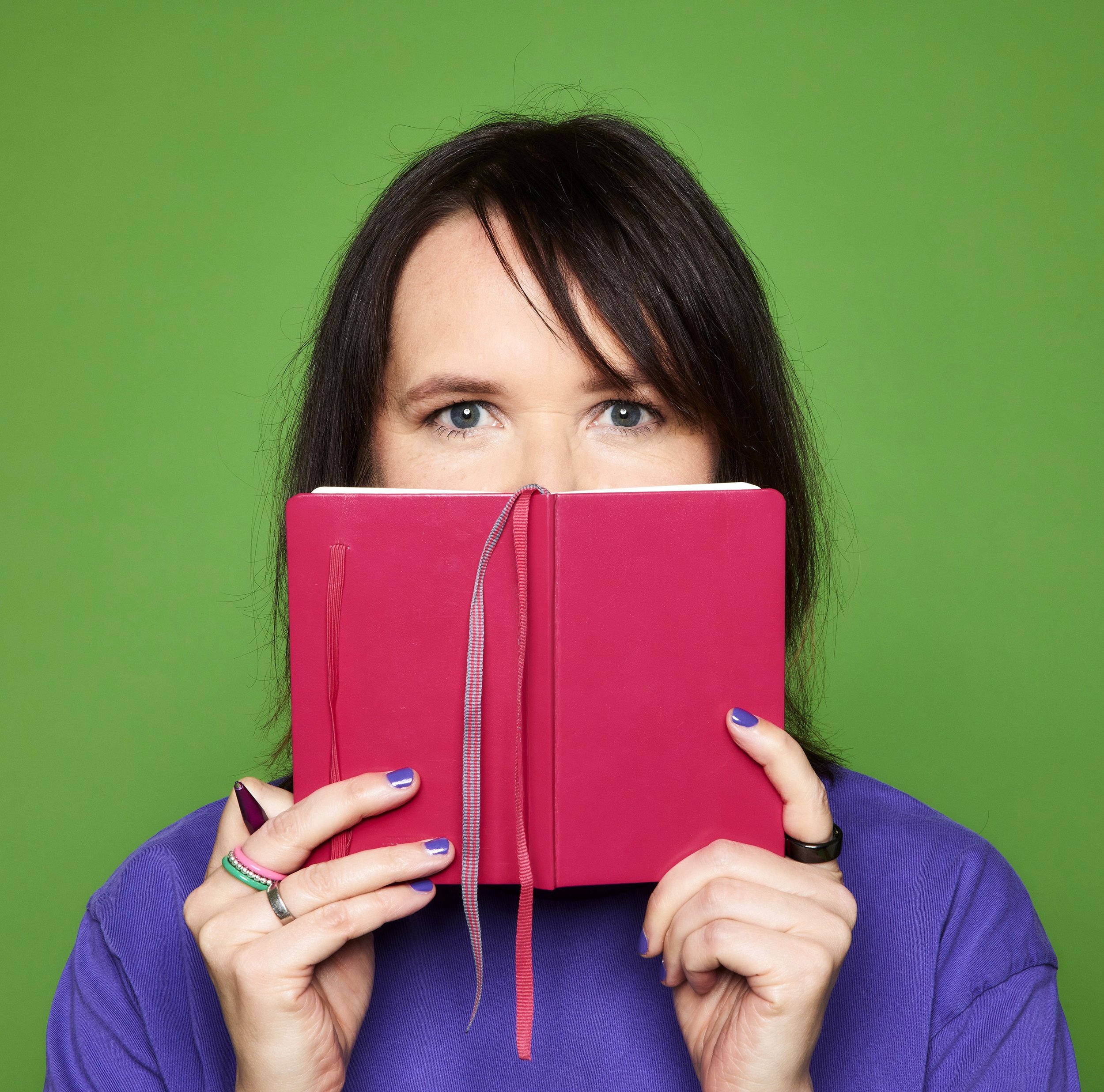 Eine Frau mit blauen Augen und schwarzen Haaren hält ein rosa Buch vor das Gesicht, so dass nur die Augen und Hände sichtbar sind, vor einem grünen Hintergrund.
