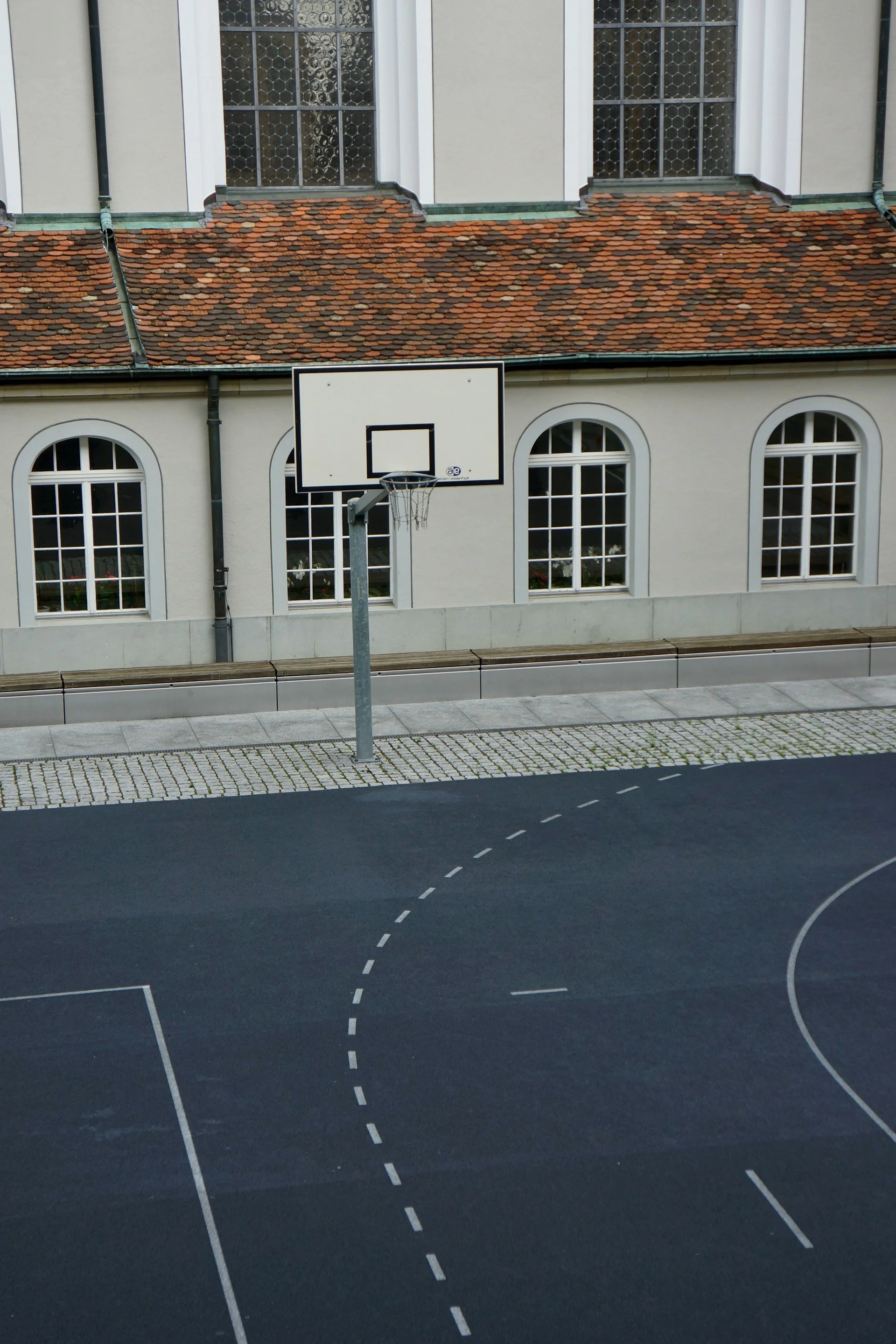 Terrain de basket extérieur avec un panier vide devant un bâtiment à fenêtres et un toit en tuiles.