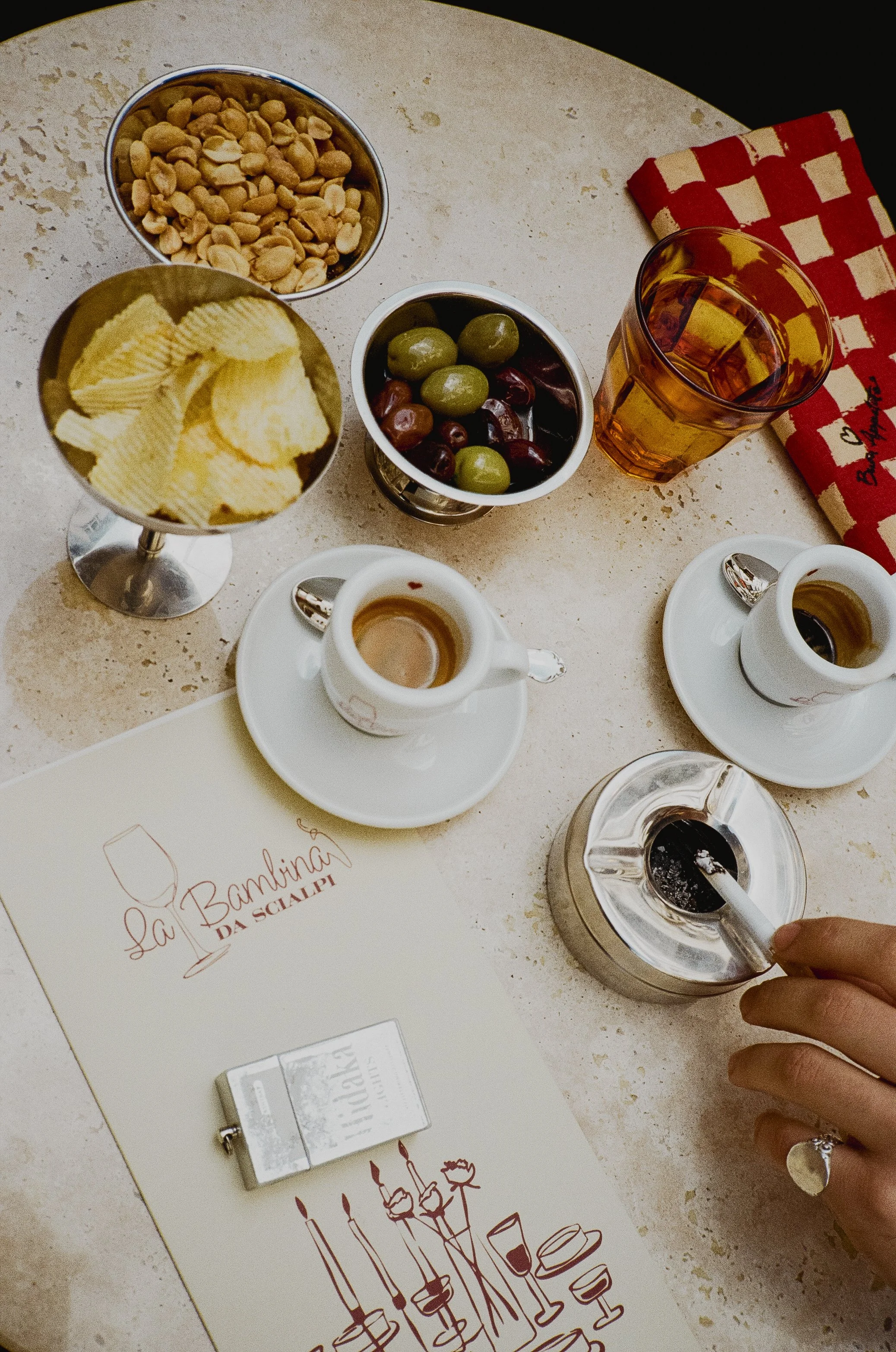 A table with snacks including potato chips, mixed olives, and peanuts, as well as coffee cups, a pink-colored drink, a napkin, and a menu for La Bambina da Sciapi.