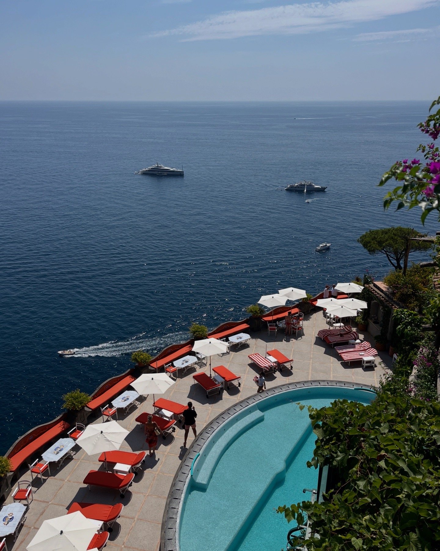 View of a swimming pool with red lounge chairs and white umbrellas overlooking the ocean with yachts and boats in the distance.