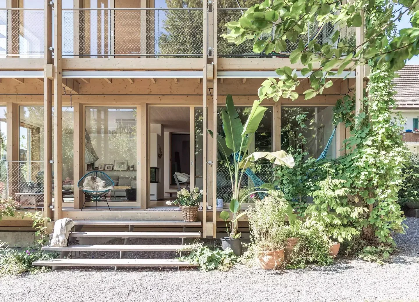 Front view of a partially constructed two-story house with a wooden frame and large glass windows. There are potted plants and greenery outside, with a gravel pathway in the foreground.