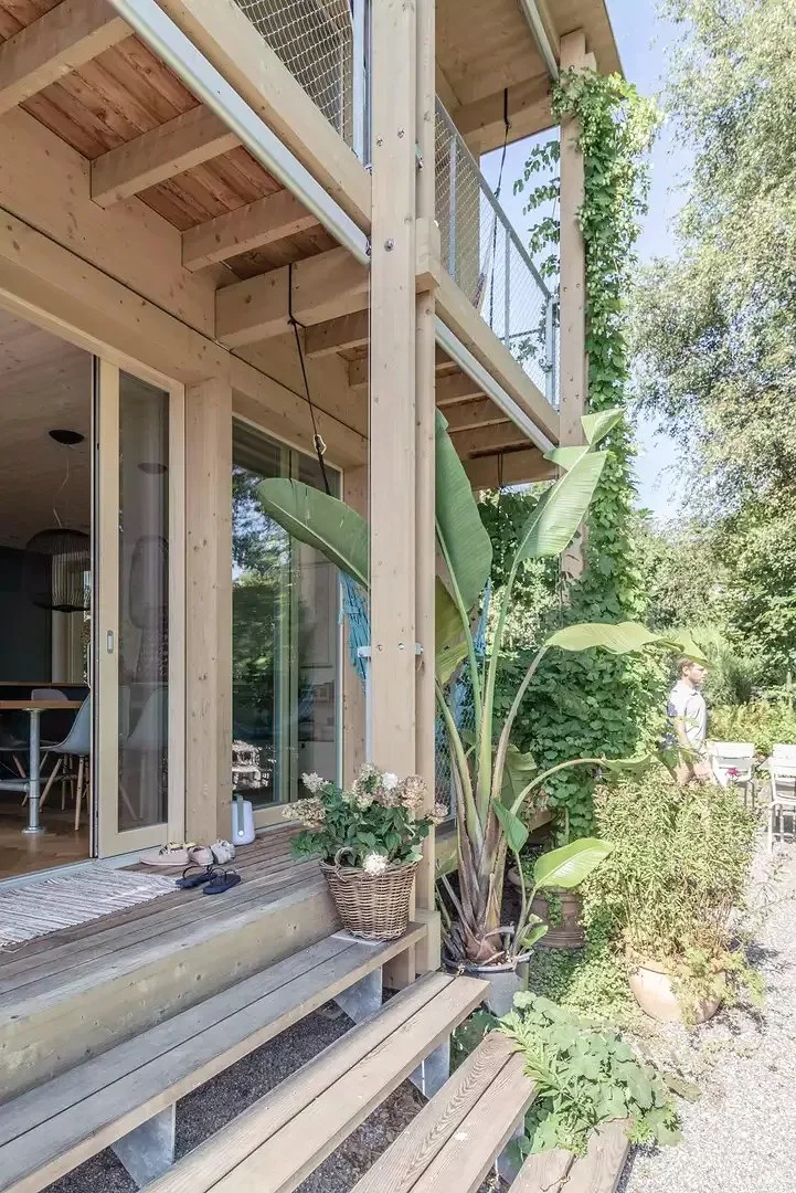 Outdoor view of a wooden deck with potted plants and stairs leading to a house with sliding glass doors, surrounded by lush greenery and trees.