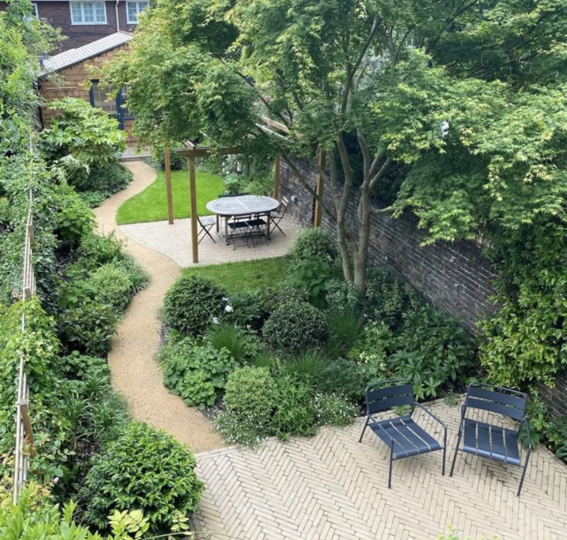 A peaceful backyard garden with lush green plants, a winding gravel pathway, a patio area with a round table and chairs, two black chairs on a brick patio, and tall trees providing shade, enclosed by a brick wall.