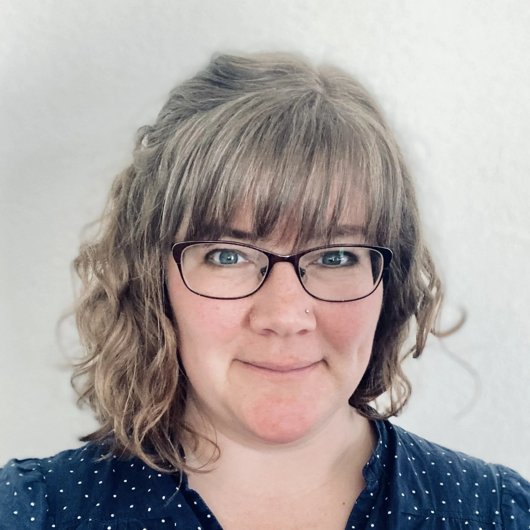 A woman with wavy, shoulder-length brown hair and glasses takes a selfie against a plain light-colored background.