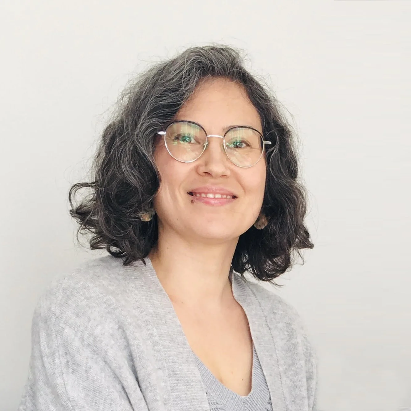 A woman with curly dark hair, wearing glasses, smiling, in front of a plain white background.
