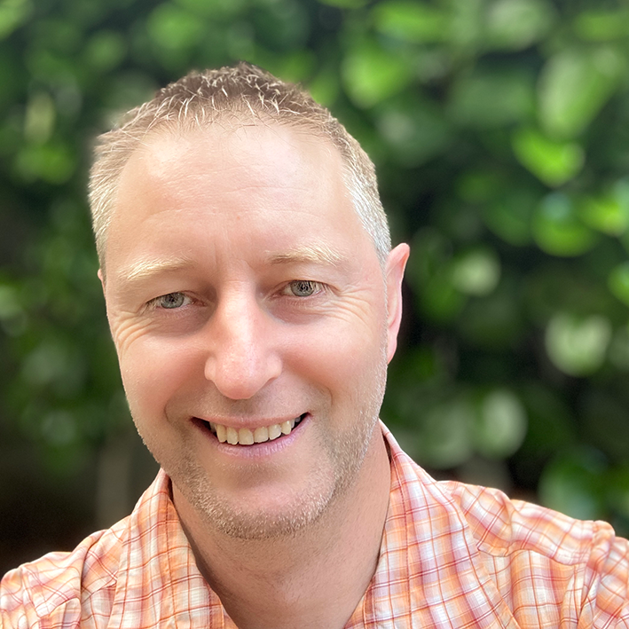 A smiling man with short hair in front of green leafy background, wearing an orange plaid shirt.