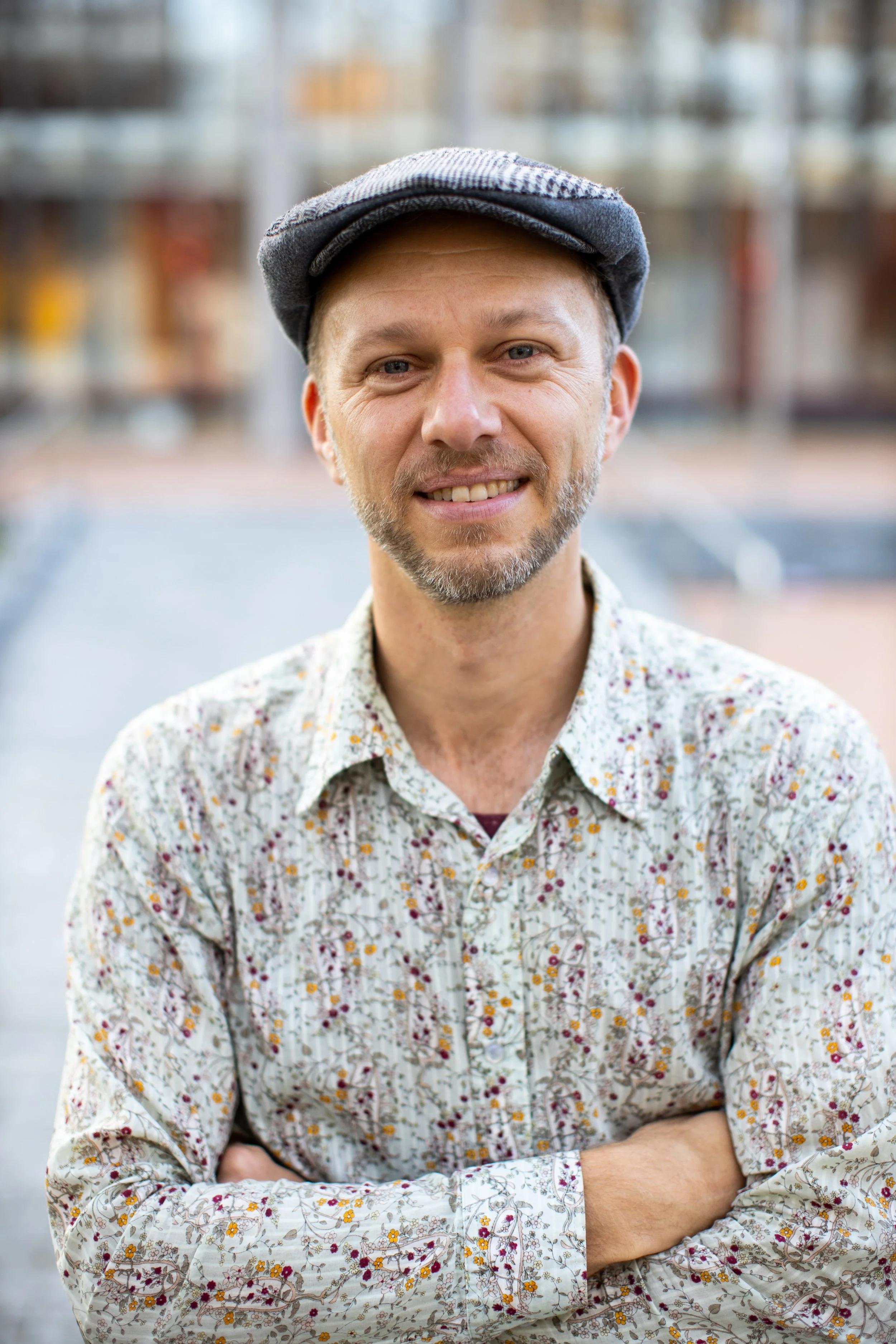 A middle-aged man with a beard, wearing a flat cap and a floral patterned shirt, smiling with arms crossed outdoors.