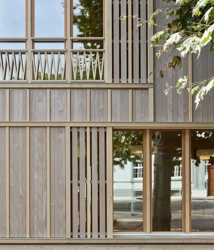 Close-up of a modern building's exterior with wooden vertical slats and large glass windows, with trees reflected in the glass.