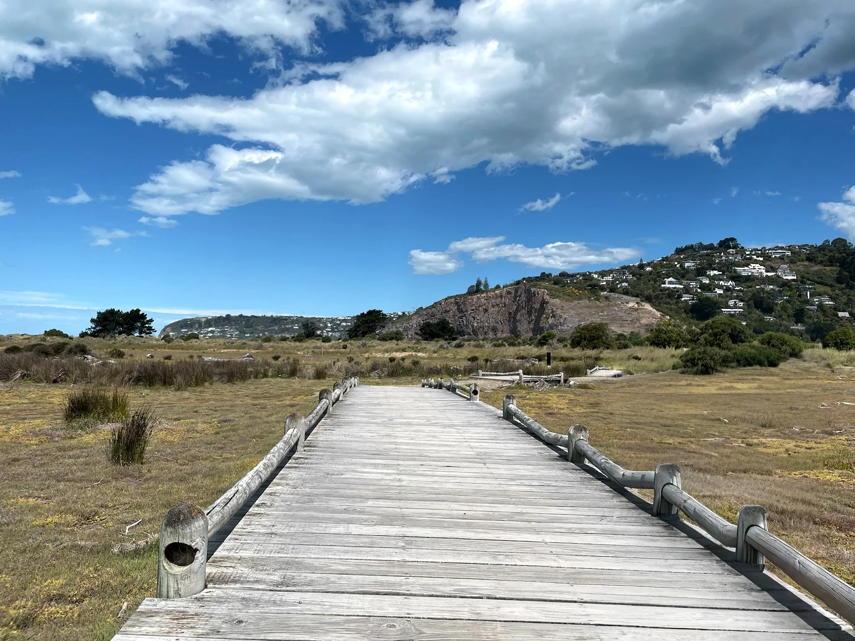 A wooden boardwalk extending through a grassy field with hills and houses in the background under a partly cloudy blue sky.