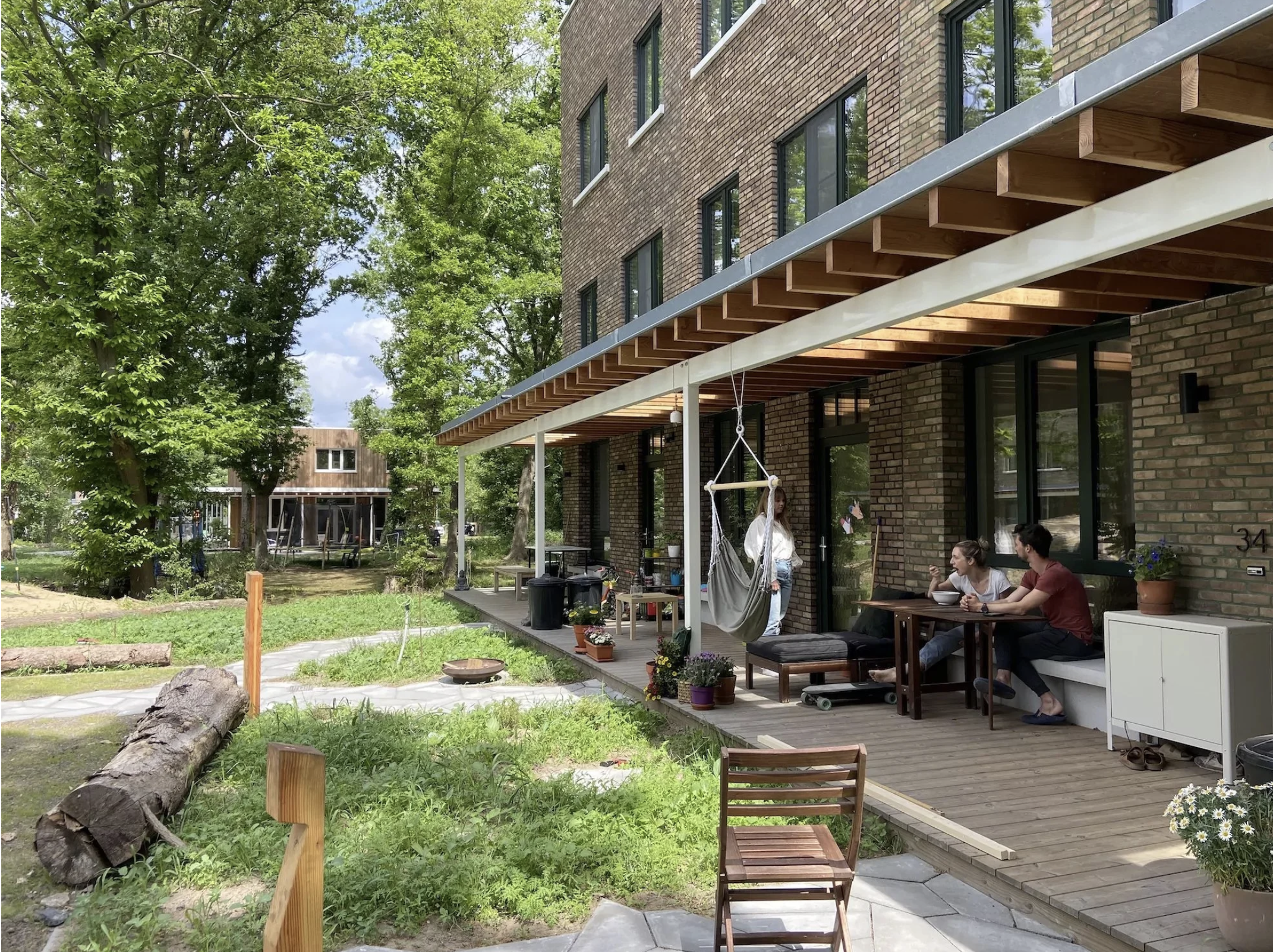 People sitting at a table on a brick apartment building's patio, with a woman standing nearby, in a green outdoor space with trees and a small playground.
