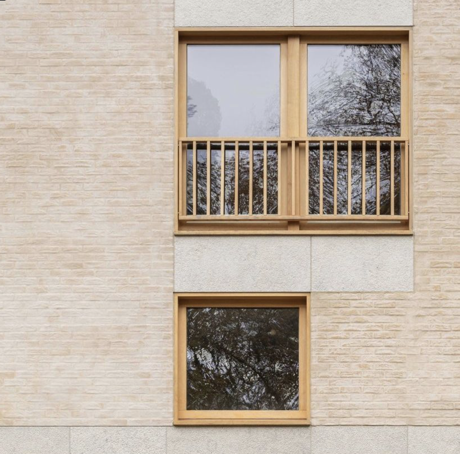 A beige brick building with three windows, one on top and two below, featuring wooden frames and a wooden balcony railing on the top window; the windows reflect trees and the sky.