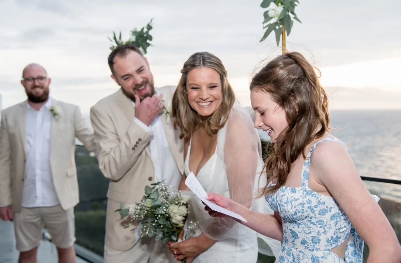 Bride and groom smiling during a wedding ceremony on a balcony with ocean view, accompanied by a person reading from a paper and another person in a light suit in the background.