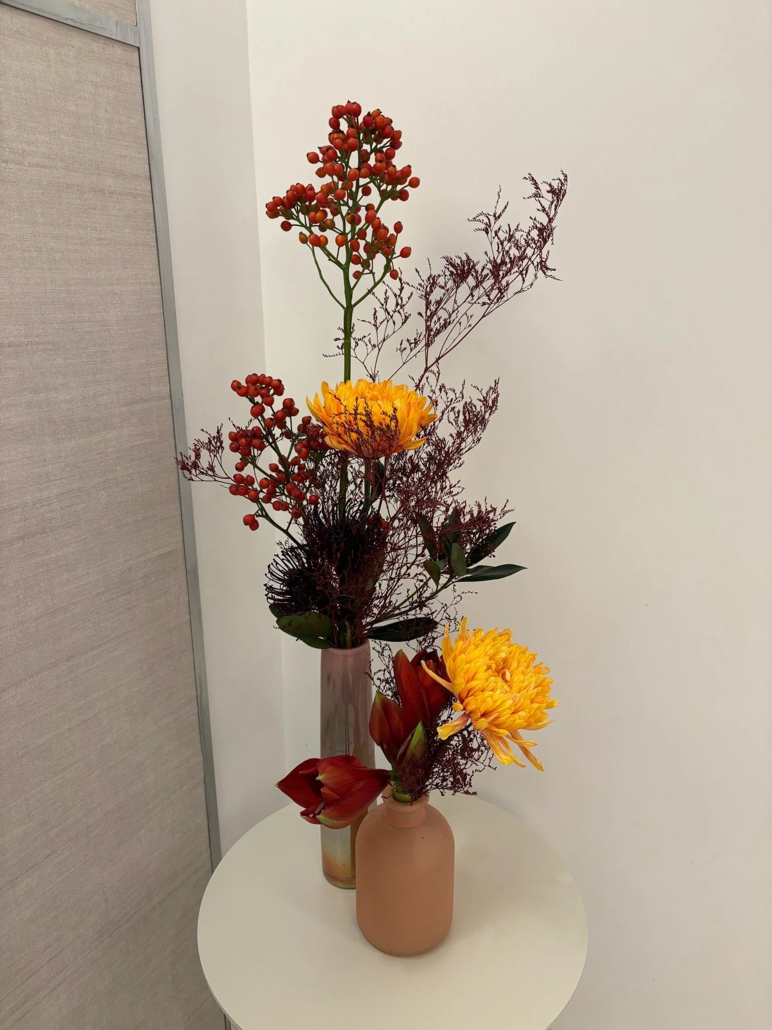 Une composition florale avec des vases en terre cuite contenant des fleurs d'automne, des branches rouges et vertes, sur une table ronde blanche, dans un décor intérieur minimaliste.