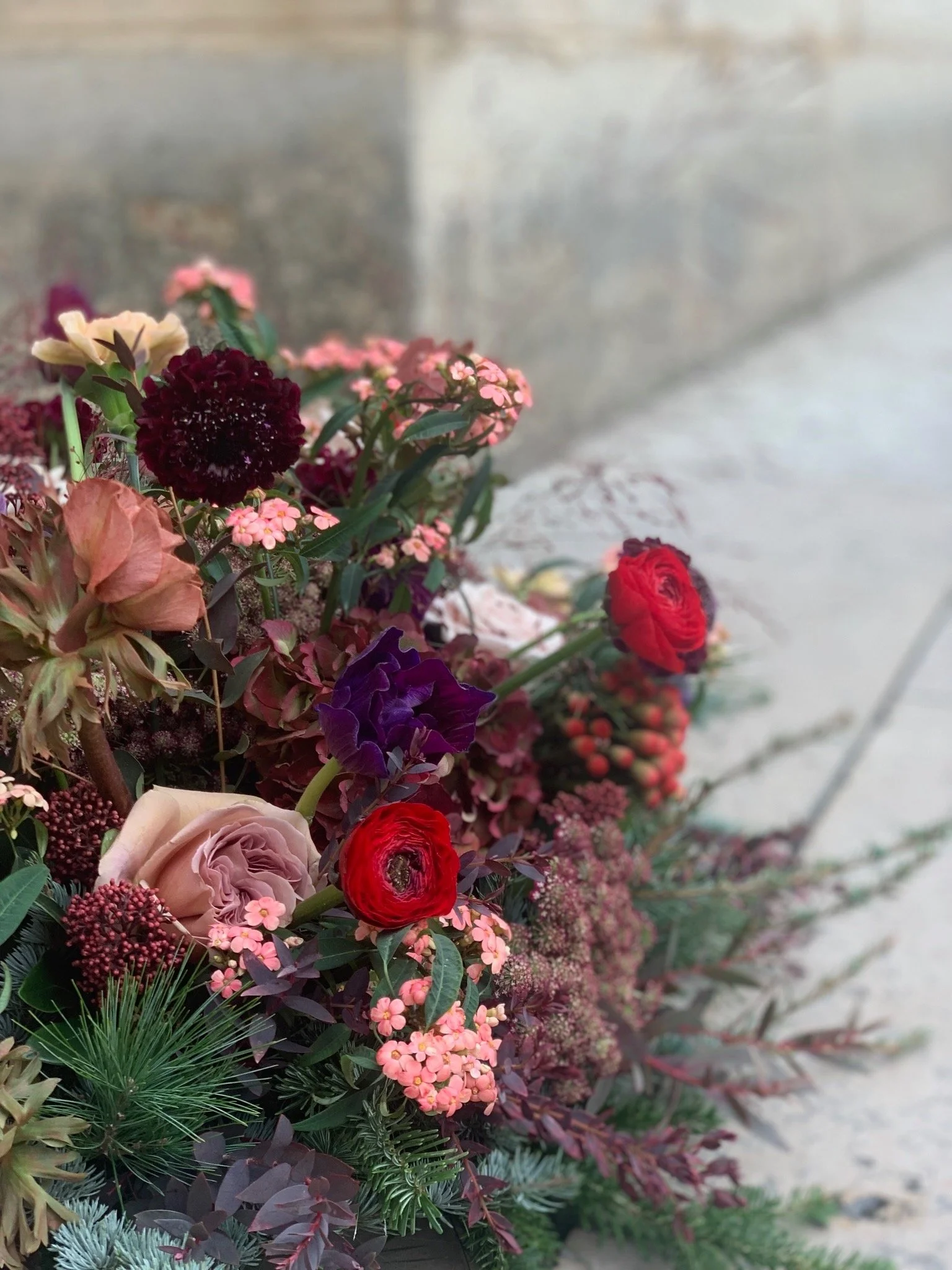 Un arrangement floral composé de roses, pivoines, petites fleurs roses, feuillage vert foncé et branches de conifère, placé devant un mur en pierre.