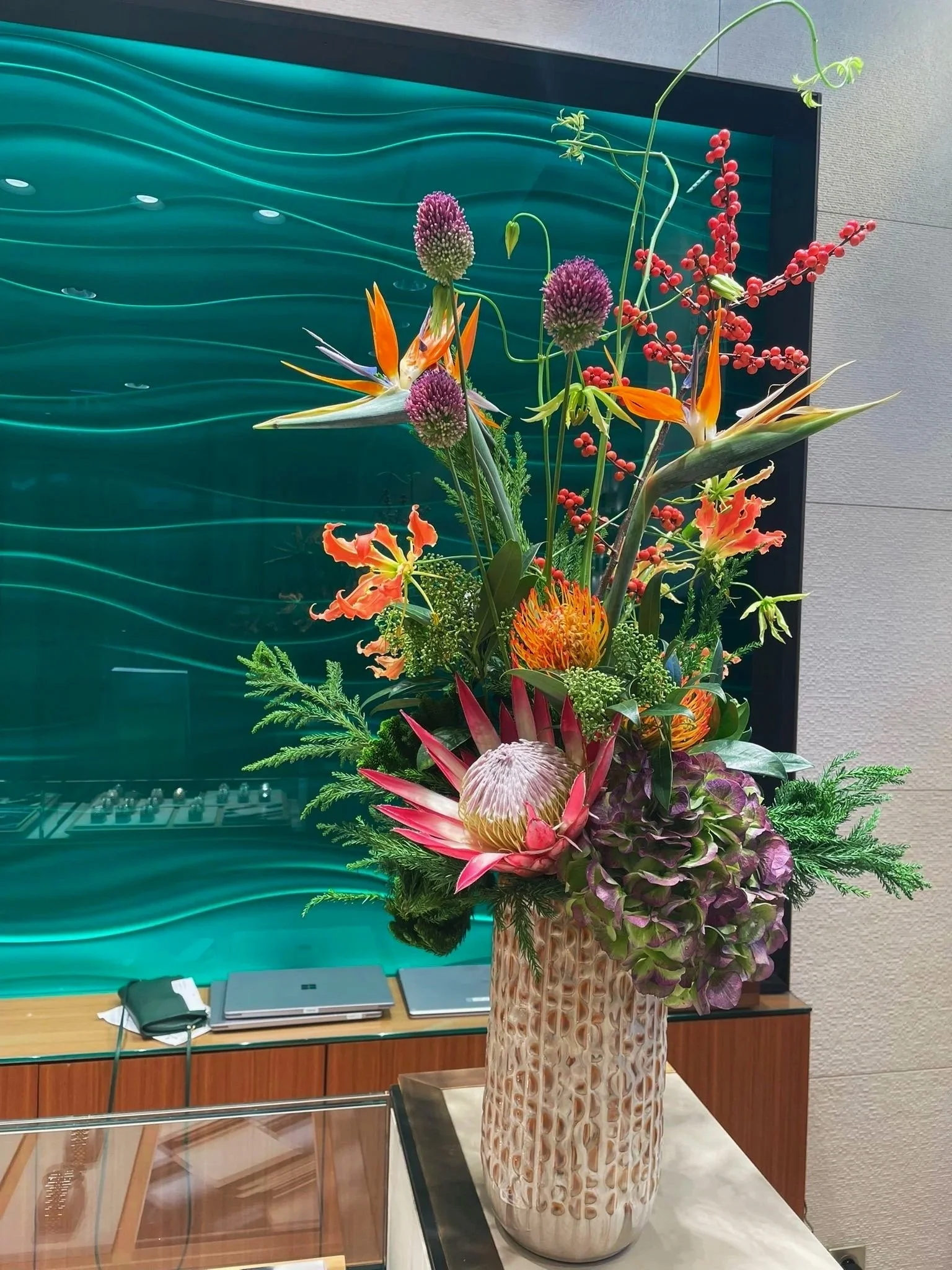 Un arrangement floral coloré dans un vase en céramique avec des fleurs tropicales et des feuillages décoratif. En arrière-plan, un mur bleu avec des vaguelettes stylisées.
