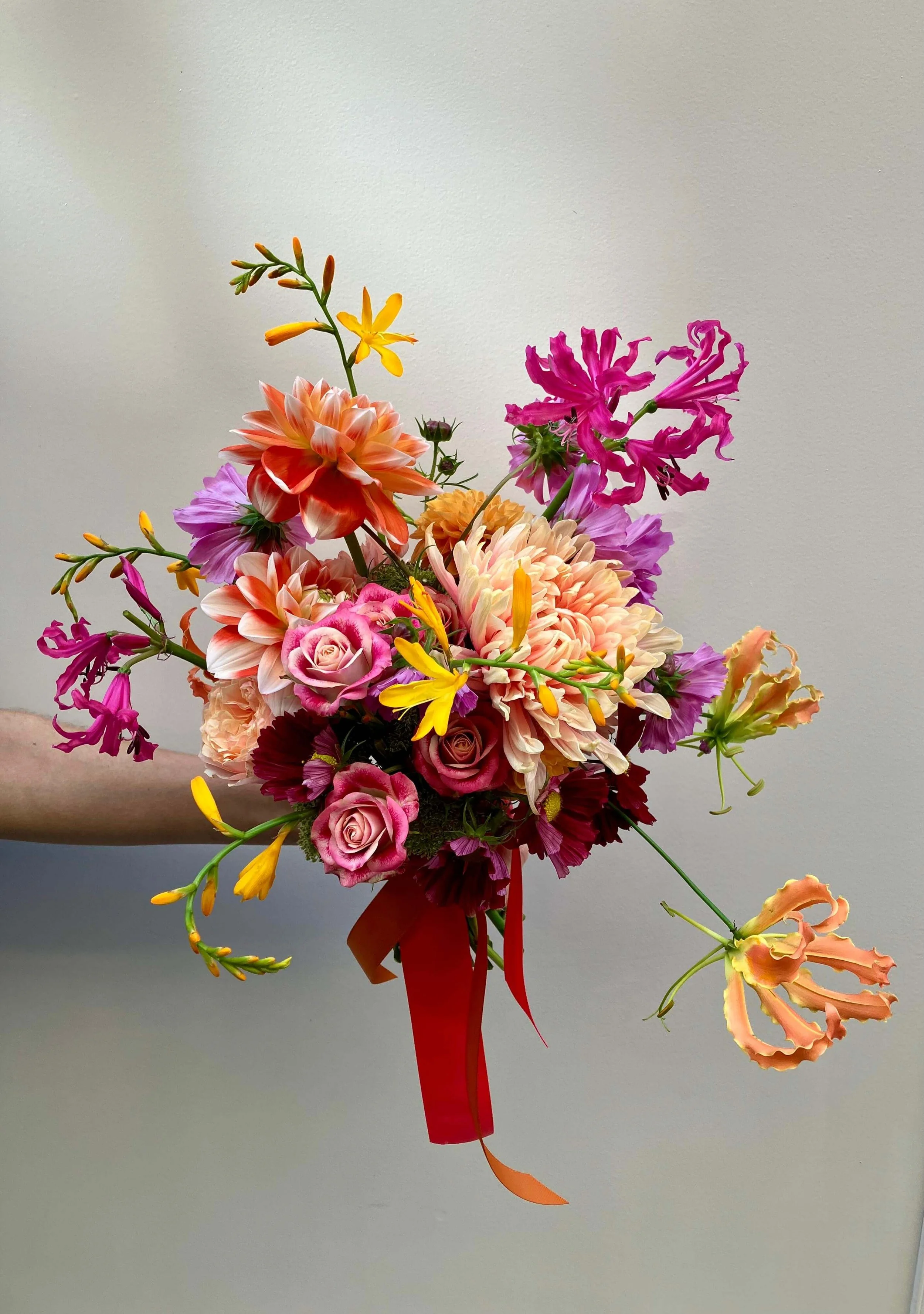 Bouquet de fleurs colorées comprenant des roses, des dahlias, des glédioles, des géorries, et d'autres fleurs, attaché avec un ruban rouge.