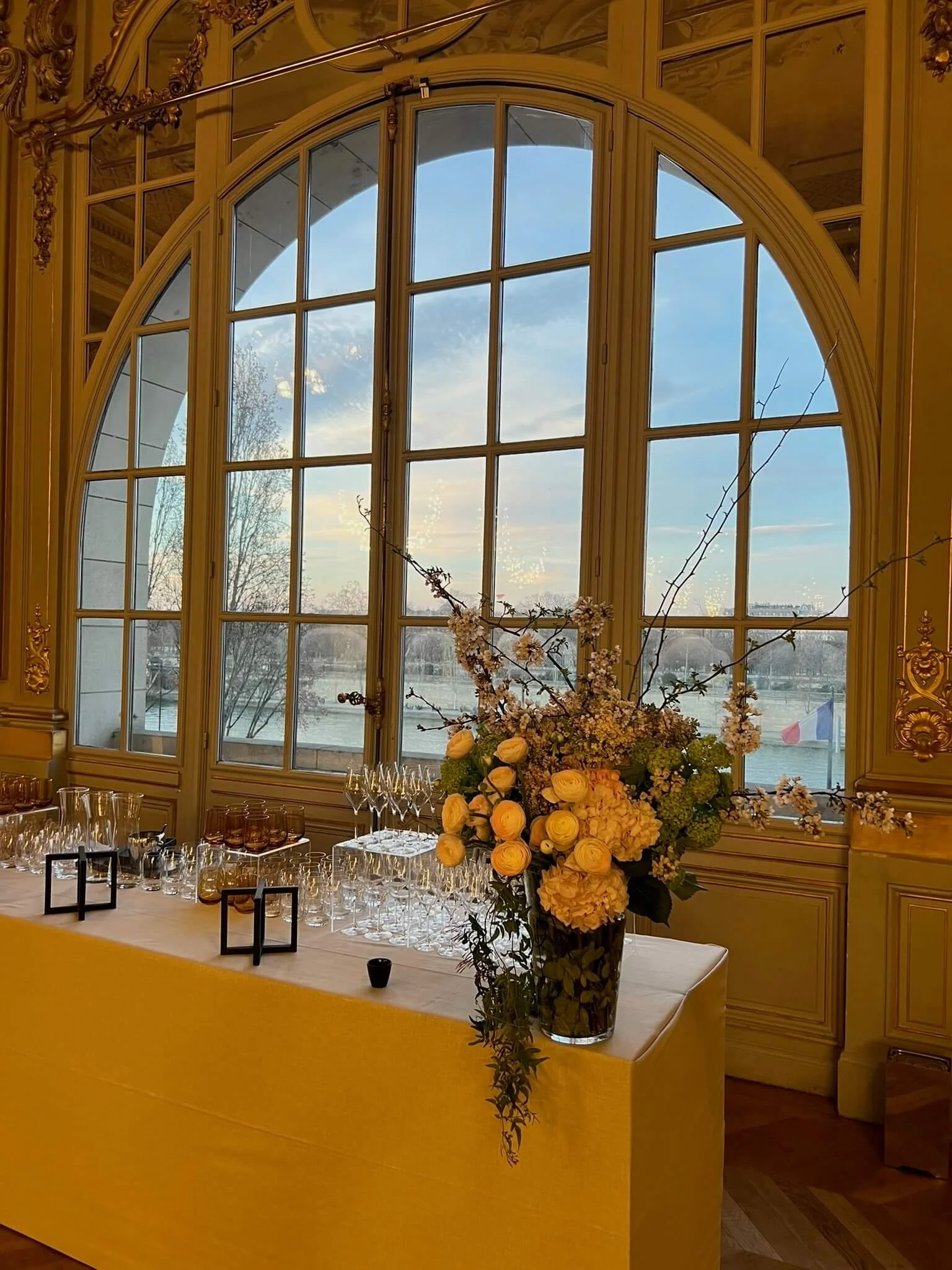 Une grande fenêtre avec des volets en bois doré offrant une vue sur une rivière et des arbres. Devant la fenêtre, un arrangement floral avec des roses jaunes, des pivoines et des branches fleuries est posé sur une table jaune, avec de la vaisselle et