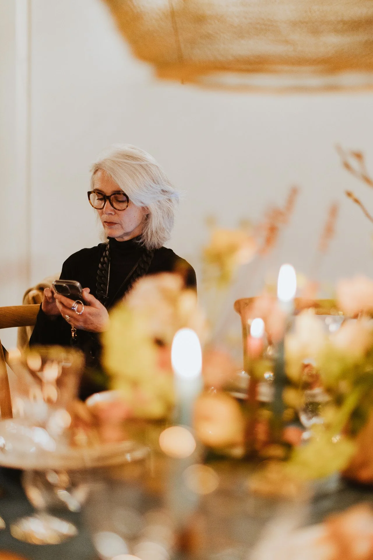 Une femme avec des cheveux gris, portant des lunettes noires et un pull noir, utilise son téléphone en s'asseyant à une table décorée avec des bougies et des fleurs.