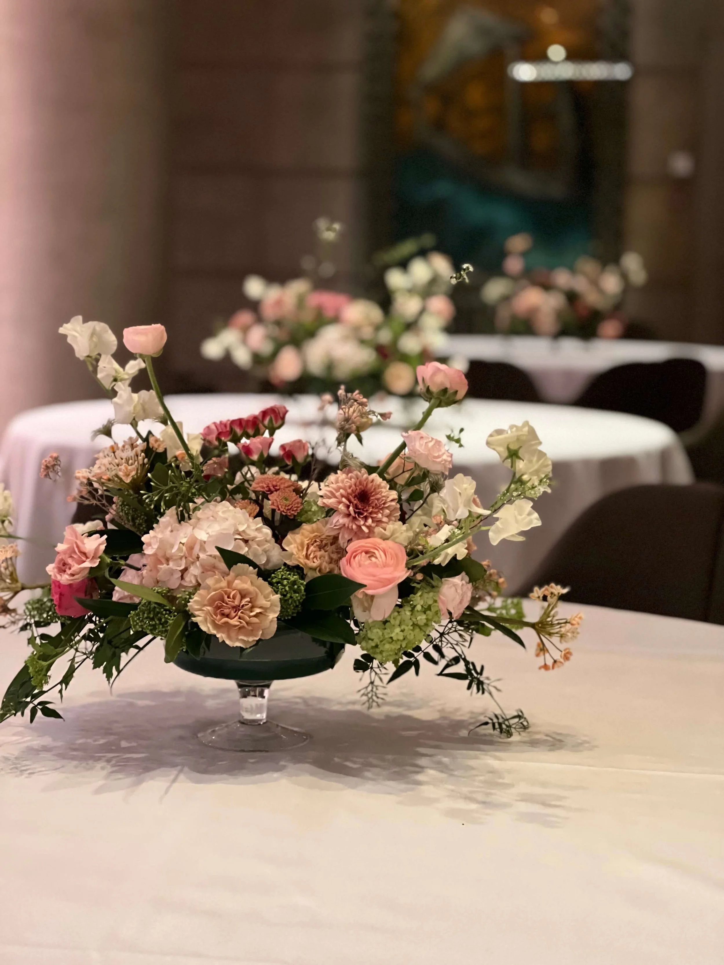 Centre de table floral avec des fleurs roses, blanches et crème sur une nappe blanche dans une salle de réception ou de banquet.