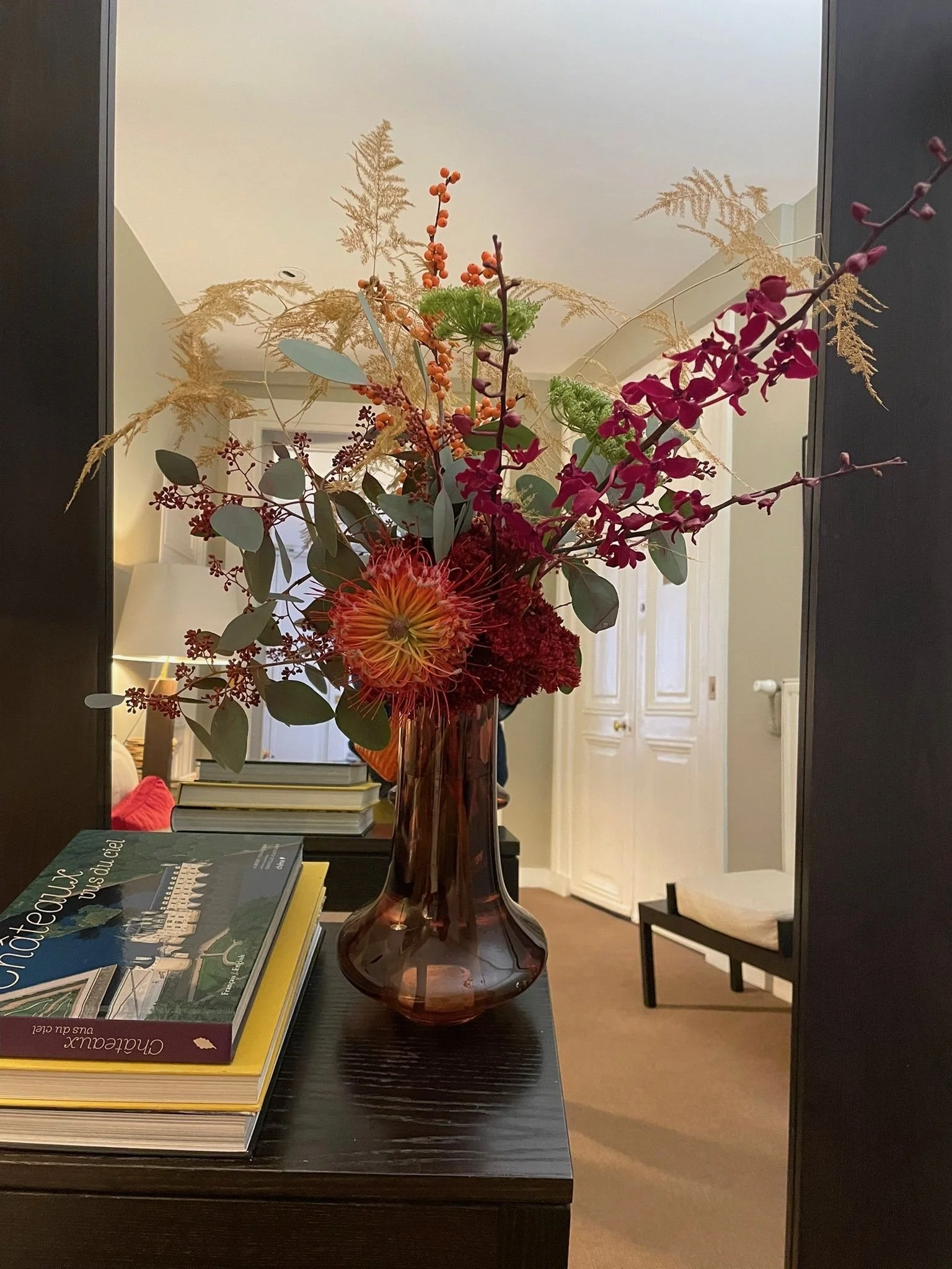 Un bouquet de fleurs colorées dans un vase en verre marron clair sur une console noire, avec des livres à côté, dans une pièce lumineuse avec un miroir derrière.