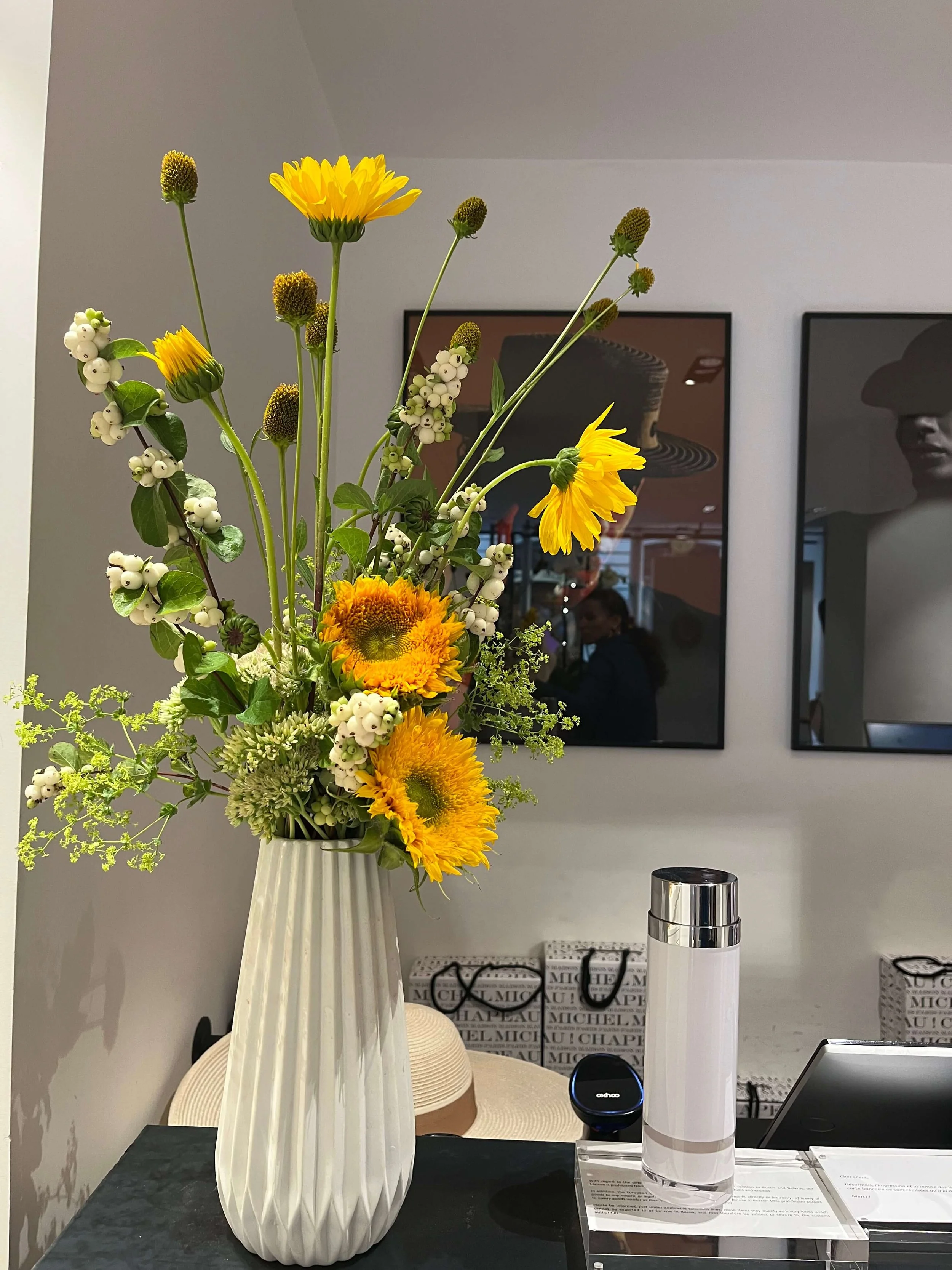 Vase blanc avec bouquet de fleurs comprenant des tournesols, des fleurs blanches et des tiges avec des bourgeons dans un intérieur moderne.