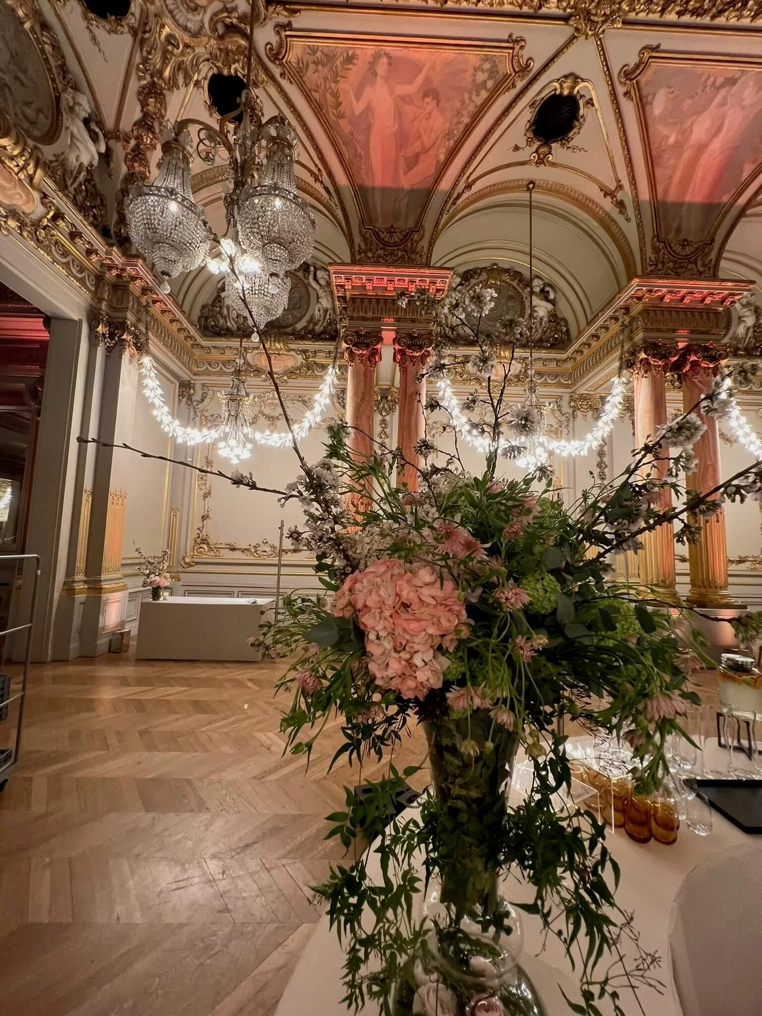 Un grand vase transparent avec un bouquet de fleurs roses, blanches et vertes, placé sur une table blanche dans une pièce ornée de moulures dorées et de colonnes aux chapiteaux corinthiens, avec un plafond décoratif et un chandelier en cristal suspen