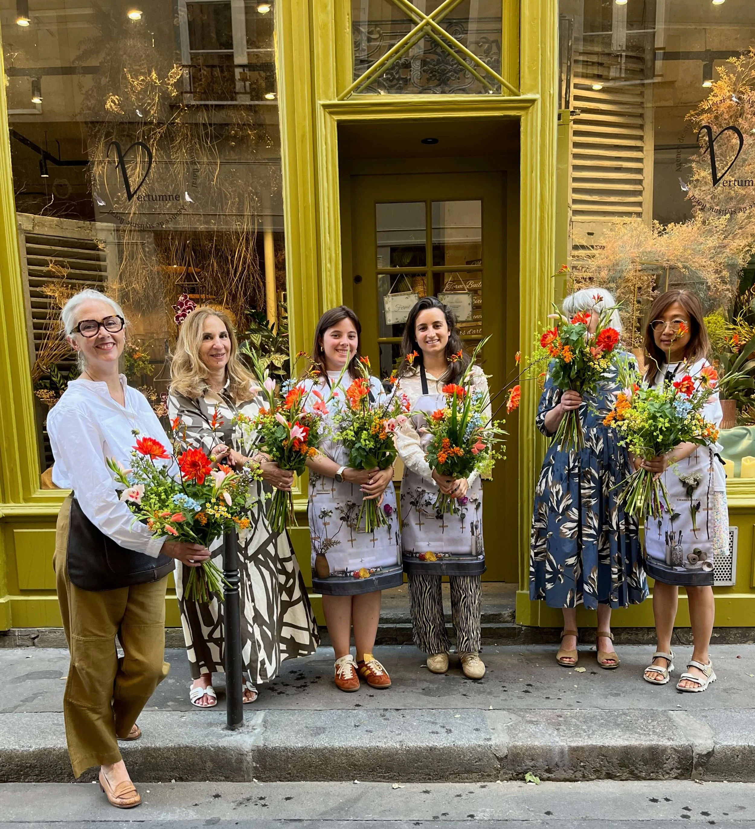 Six femmes debout devant une vitrine, tenant des bouquets de fleurs colorées, souriantes, posant pour une photo.