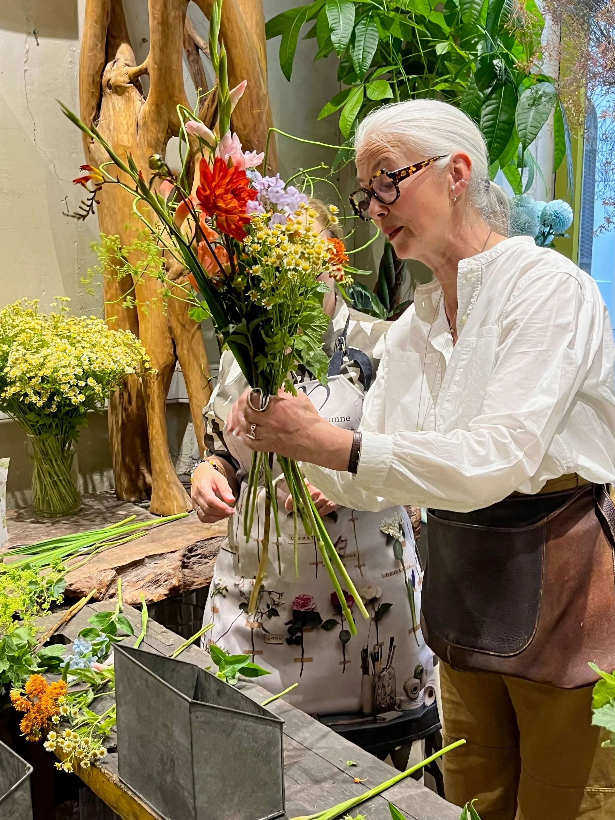Une femme, avec des cheveux gris, porte des lunettes et une chemise blanche, tenant un bouquet de fleurs colorées dans un atelier floral.