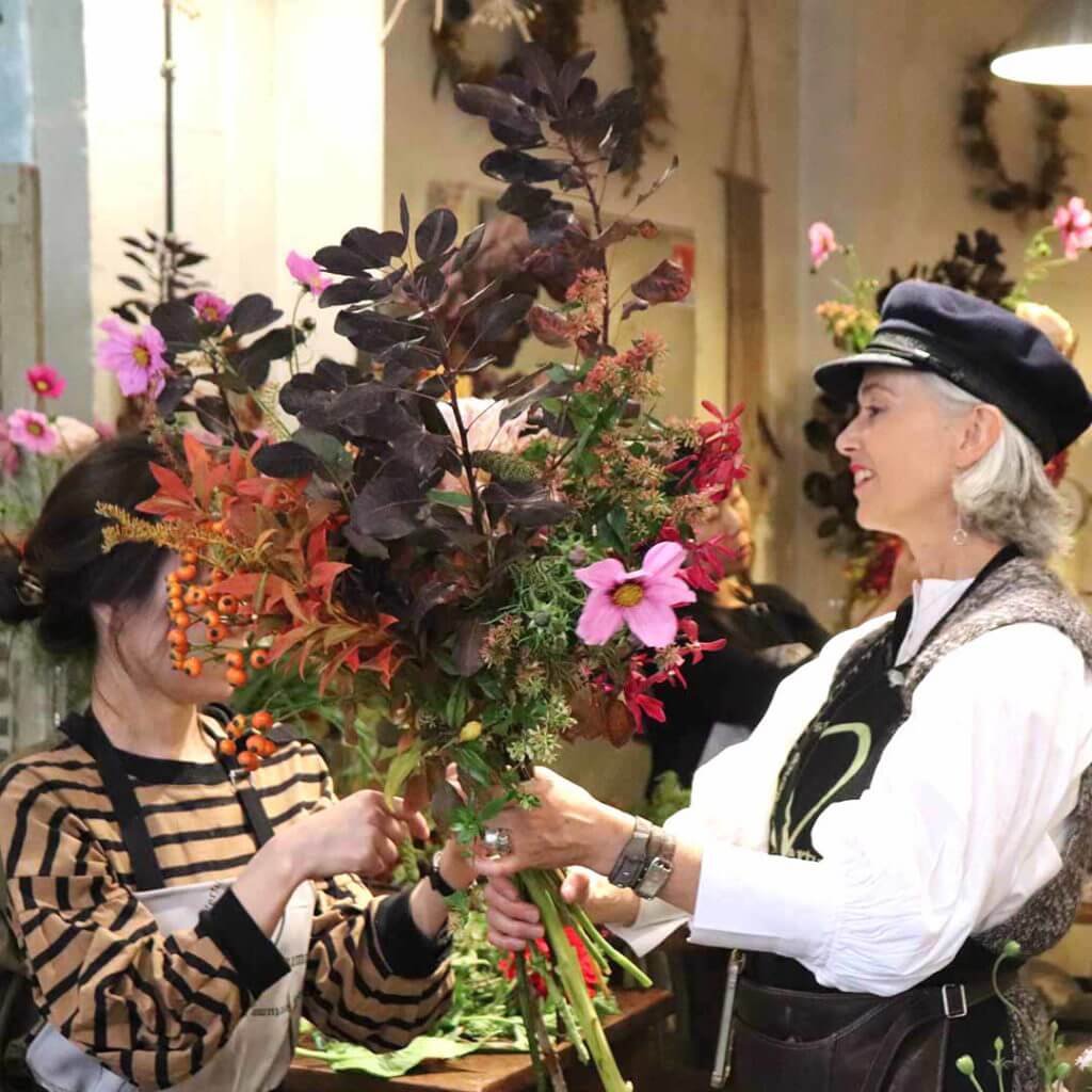 Scène conviviale en atelier autour d’un bouquet aux teintes chaudes. Partage et apprentissage dans une ambiance joyeuse.