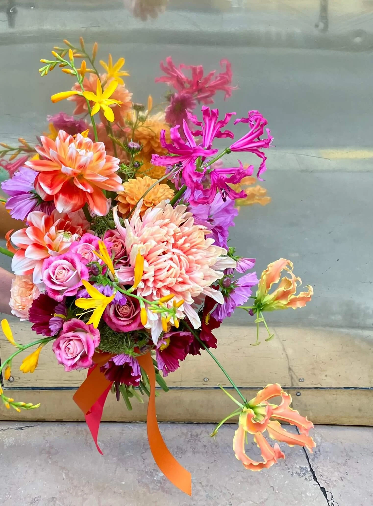 Un bouquet de fleurs colorées avec des roses roses, des dahlias orange, des iris roses et orange, des lis jaunes et roses, et d'autres fleurs multicolores, avec un ruban orange.