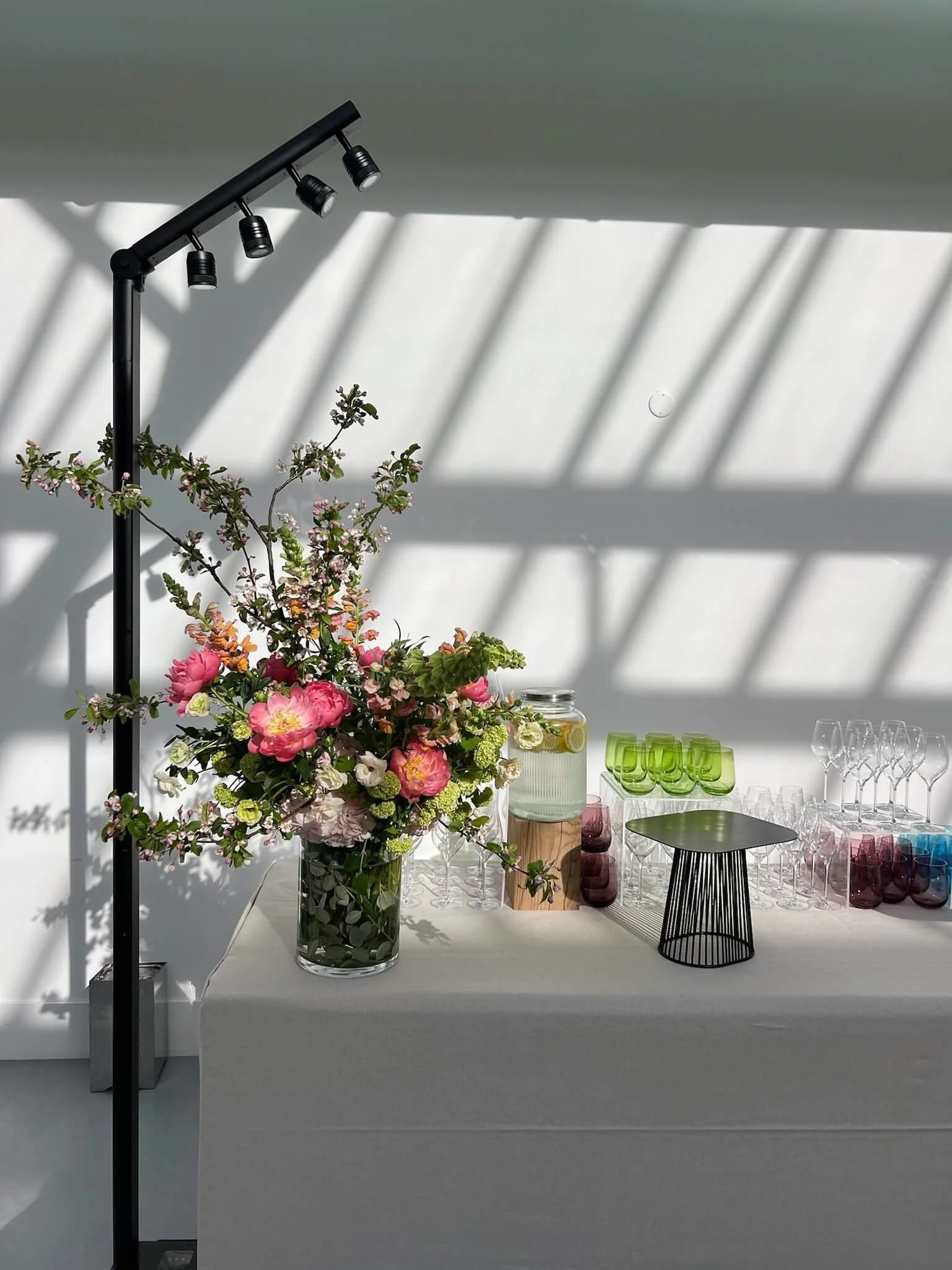 Un arrangement floral avec des fleurs roses, blanches et vertes sur une table, entourée de verres colorés, de petits plateaux, et d'une lanterne noire, avec des ombres de barres en biais projetées sur le mur et le plafond, créant un effet de lumière 