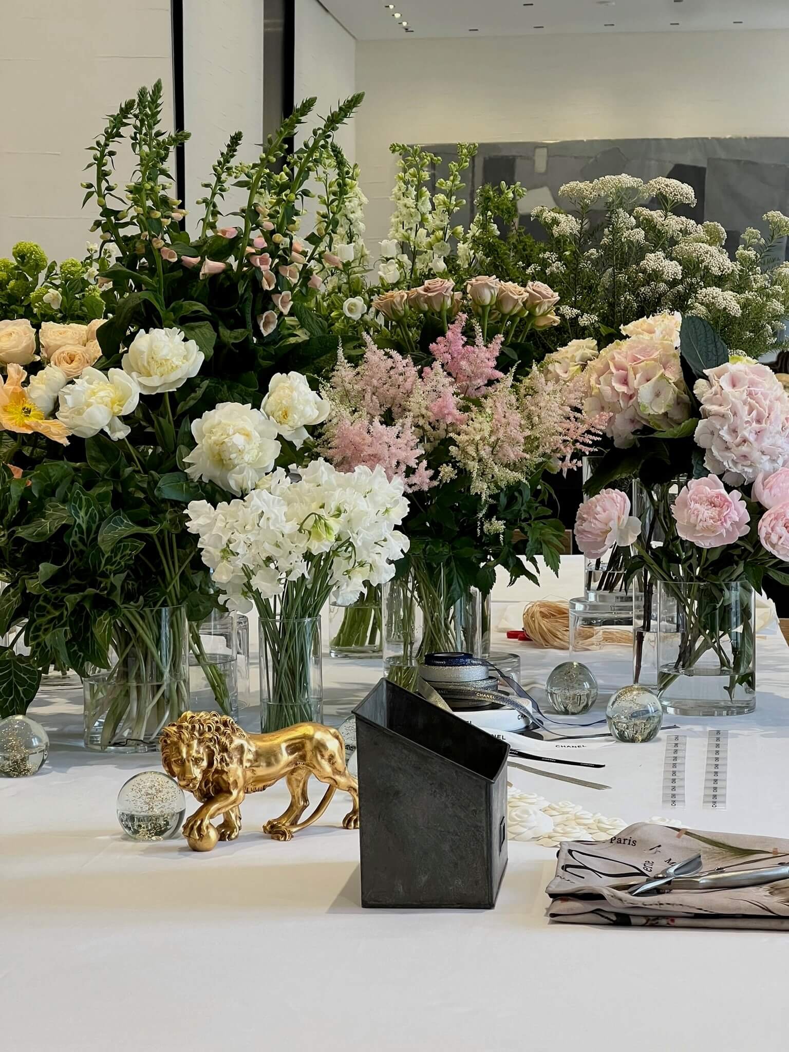 Une table avec plusieurs bouquets de fleurs dans des vases, une figurine de lion dorée, un petit objet noir, des papiers, des balles en verre et d'autres décorations.