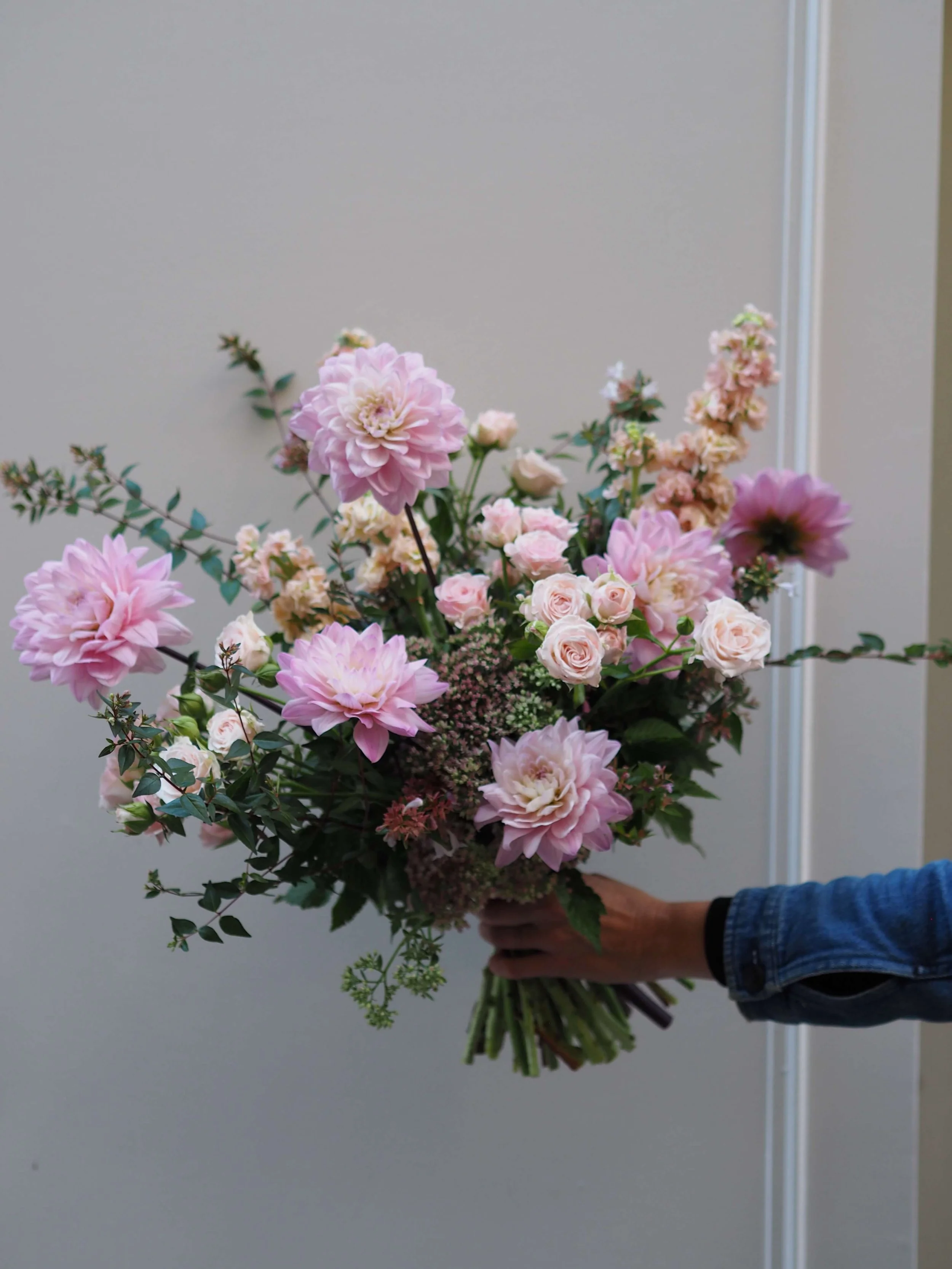 Un bouquet de fleurs roses et blanches, comprenant des dahlias, roses et autres fleurs, tenu par une personne portant une veste en jean.