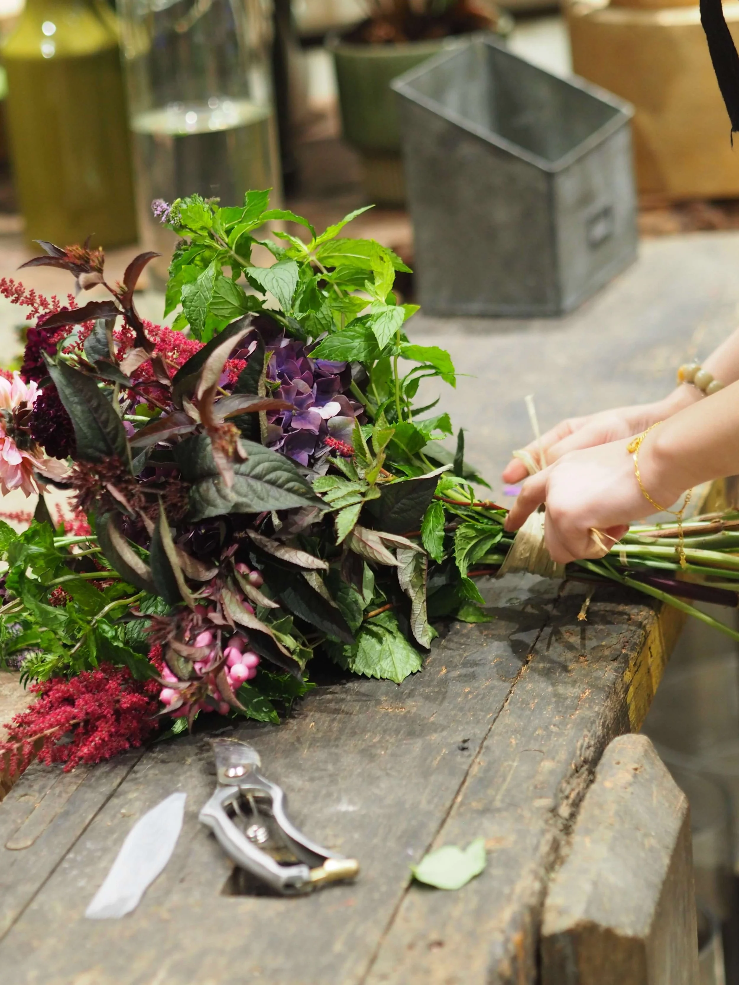 Les mains d'une personne préparant un bouquet de fleurs colorées sur une table en bois, avec outils de coupe à proximité.