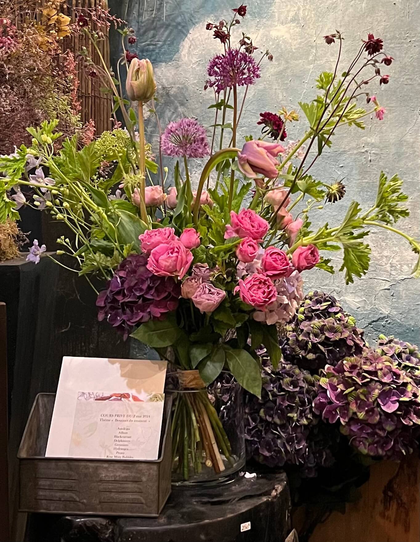 Bouquet structuré aux teintes mauves et roses, mis en scène sur un fond artistique. Élégance et mouvement floral.